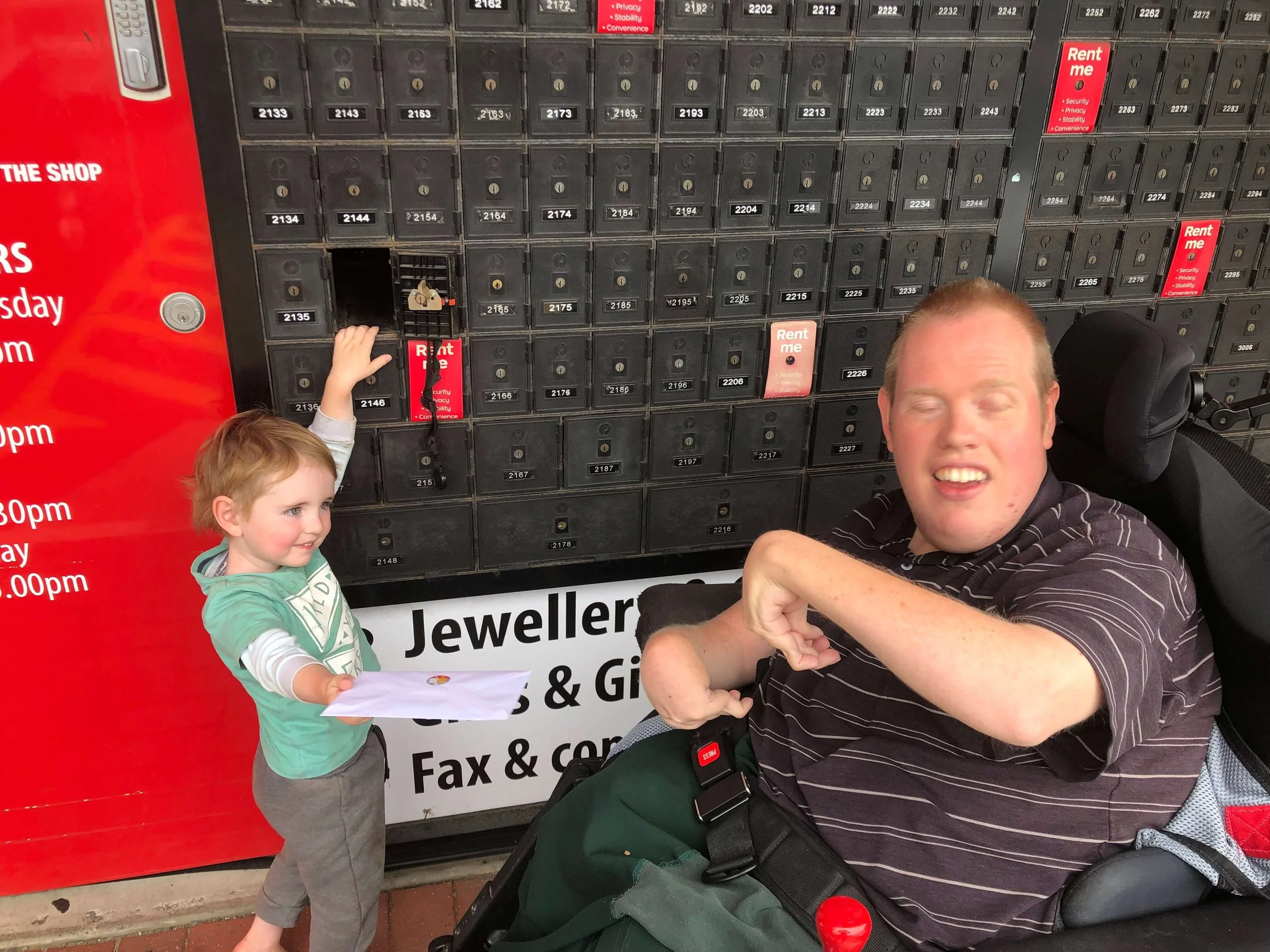 A young boy in a green shirt and gray pants holding an envelope standing next to a large black mailbox with many small lockers. An adult man in a wheelchair, wearing a black striped shirt, is smiling with his eyes closed. The background shows a red s