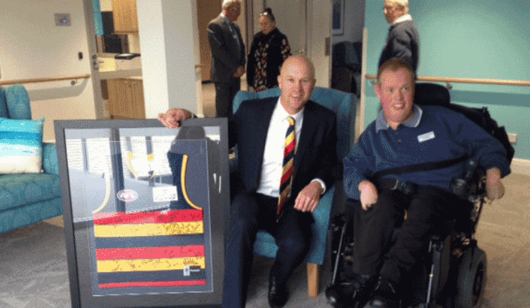 Two men, one in a wheelchair, smiling and holding a framed collage or display behind a seated man in a blue armchair. Four people are in the background, engaged in conversation.