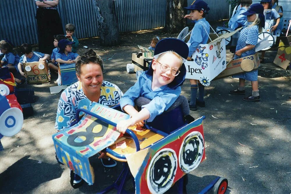 Children participating in a parade or event, riding decorated cardboard cars outdoors under trees, wearing blue uniforms or costumes.