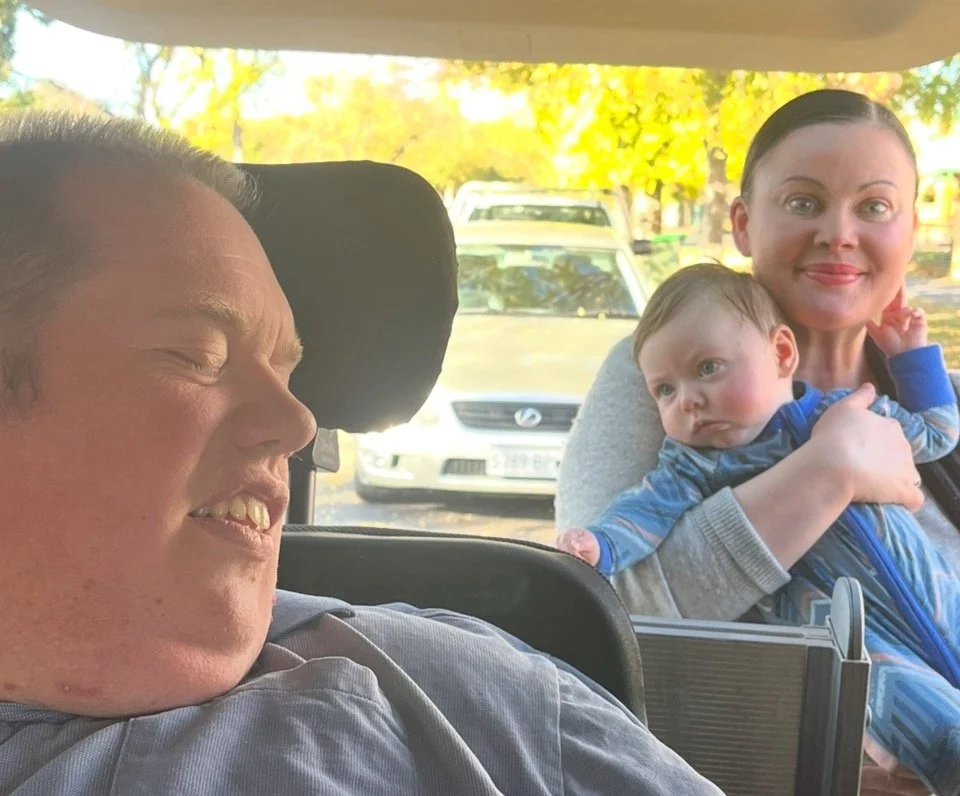 A man with Down syndrome smiling with eyes closed in a car seat, a woman holding a child with light skin and brown hair, smiling, in the background outside the car.