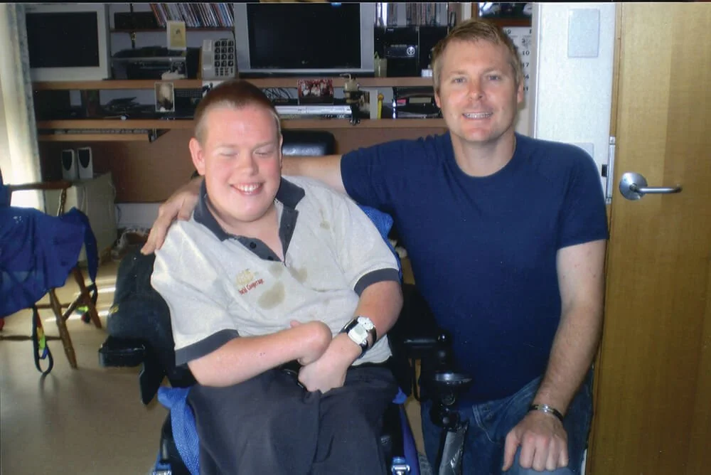 Two men smiling and sitting close together in a room with bookshelves. One man is in a wheelchair, and the other has his arm around him.