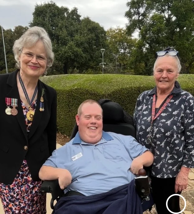 Three people outdoors: a young man in a wheelchair smiling, flanked by two older women, one with medals on her jacket and the other with glasses on her head, with trees and cloudy sky in the background.