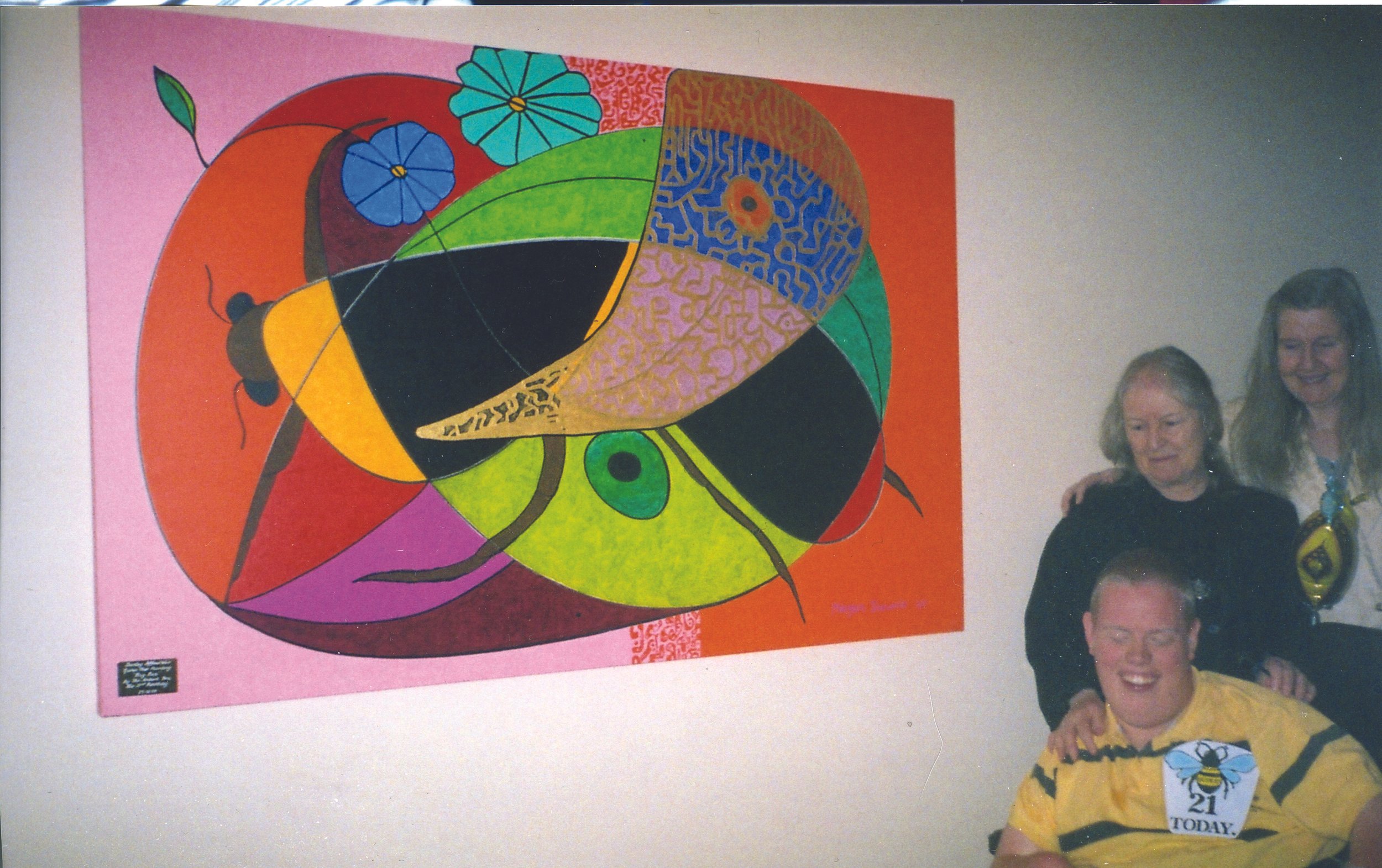 Two women and a young man in a room with a large colorful abstract butterfly painting on the wall. The young man is smiling and wearing a yellow shirt with a bee picture. One woman is standing behind him, with her hand on his shoulder, and the other 