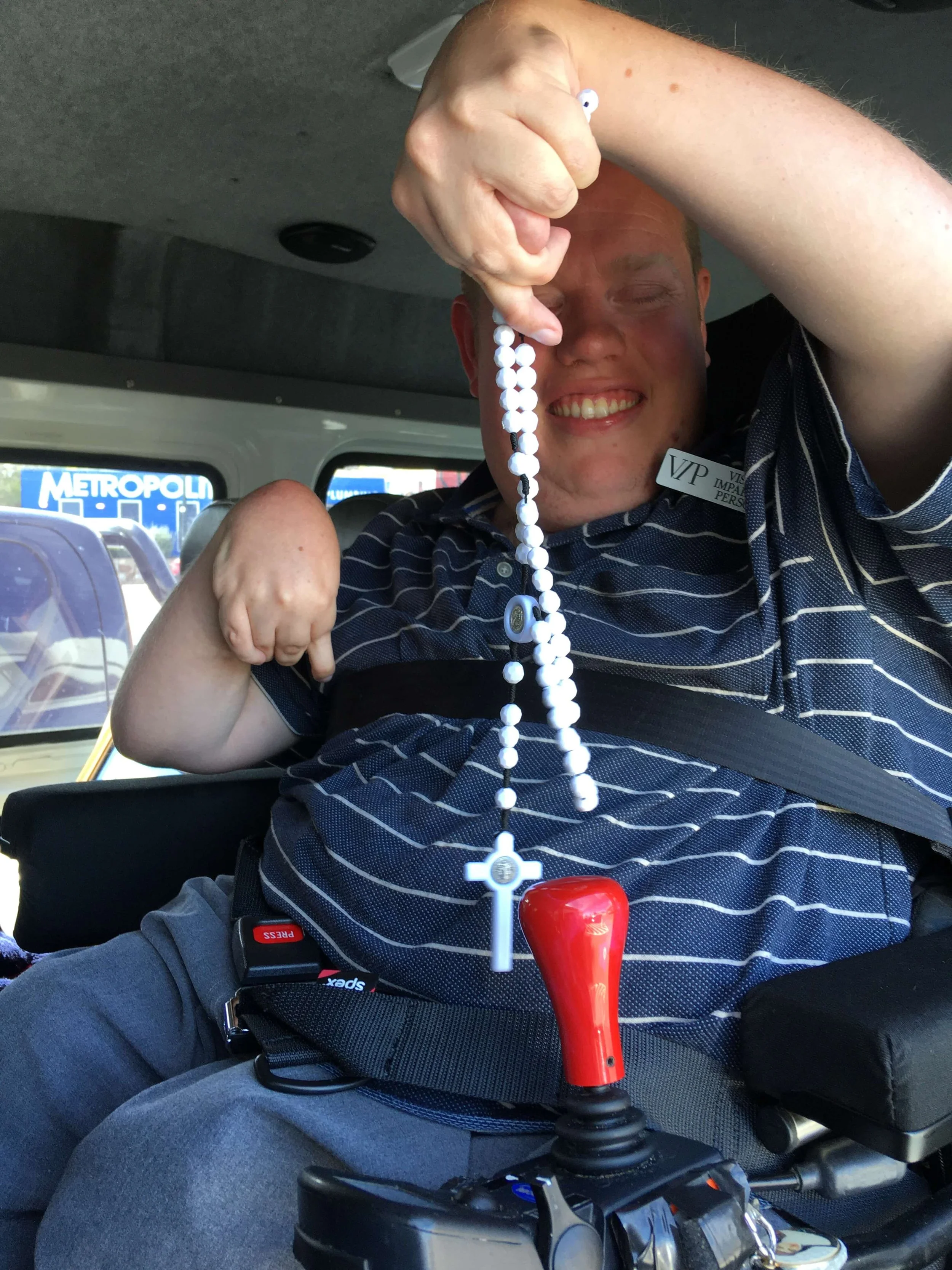 Man smiling while holding a rosary with a red bead, sitting in a wheelchair with a seatbelt, inside a vehicle with traffic outside.
