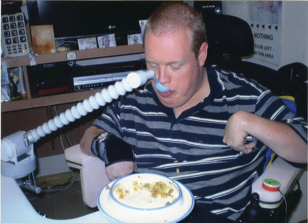 Man with a nasal oxygen tube sitting at a table with a plate of food in front of him, in a room with photos and electronics on the shelves behind him.