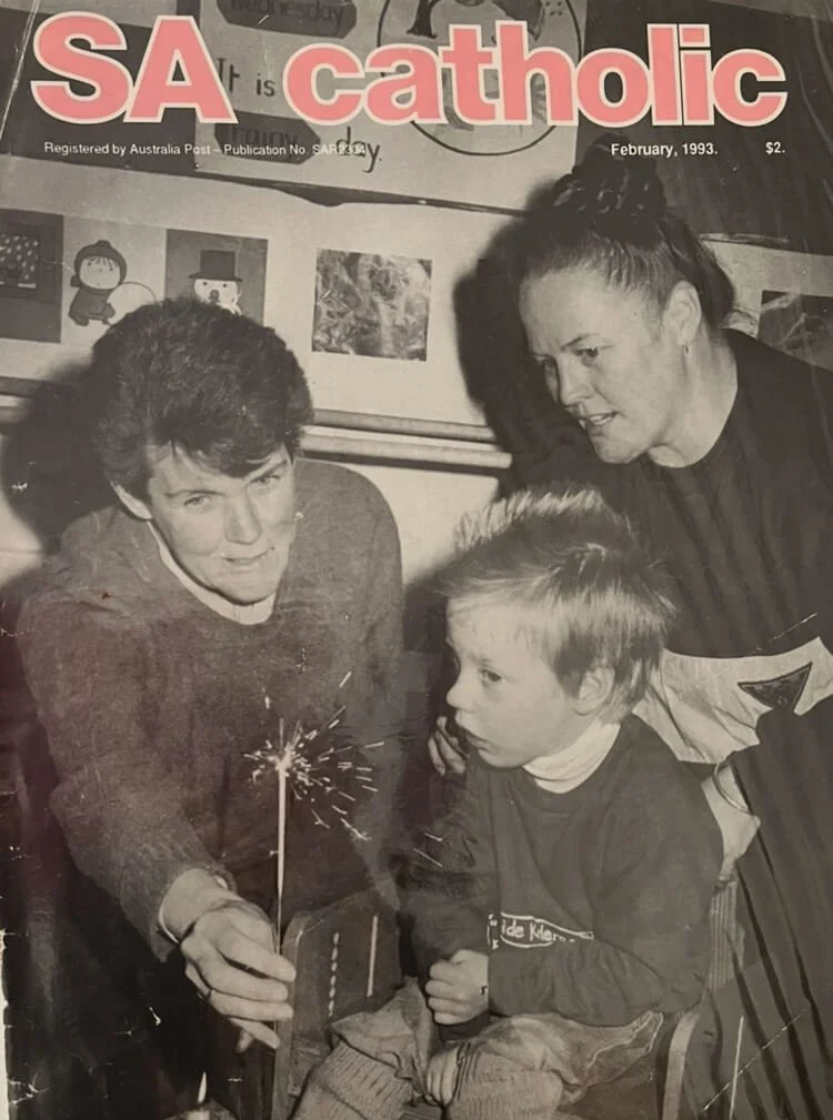 Black and white photo from a 1993 magazine cover showing two adults and a child gathered around a lit sparkler, with a poster behind them that says 'SA Catholic'.
