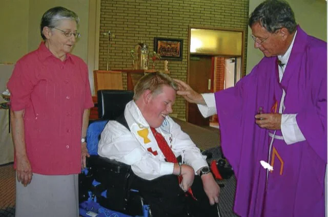 A person in a wheelchair being blessed or prayed over by a priest wearing purple vestments, with an elderly woman observing nearby.