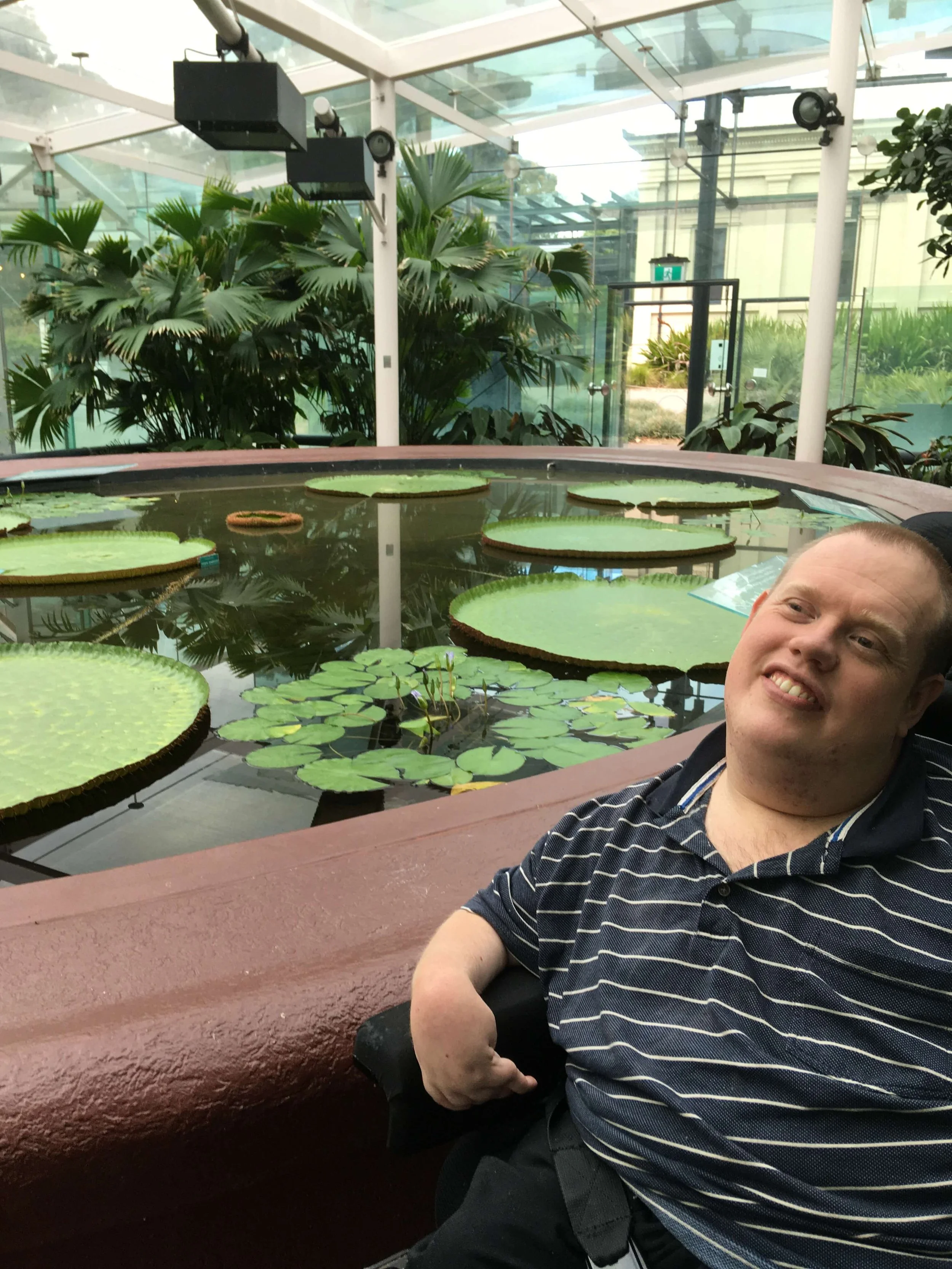 A man smiling and sitting near a water lily pond inside a greenhouse or conservatory.