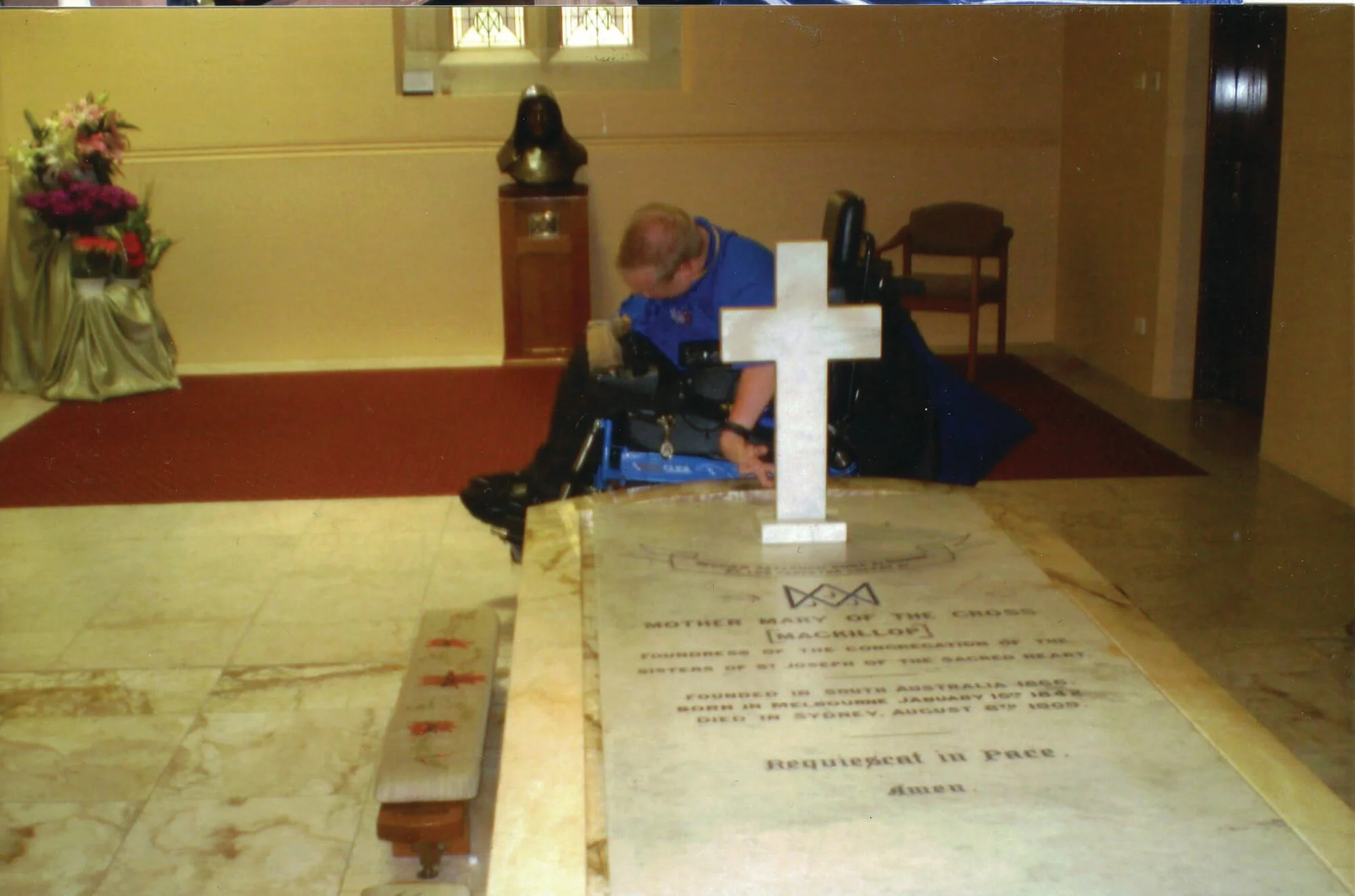 A person in a wheelchair kneeling in front of a grave with a white cross, inside a chapel with floral arrangements and a bust sculpture in the background.