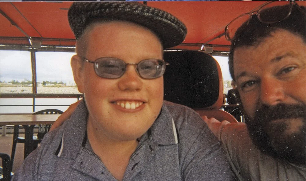 Two men taking a selfie together in an outdoor setting with a railing and water in the background.