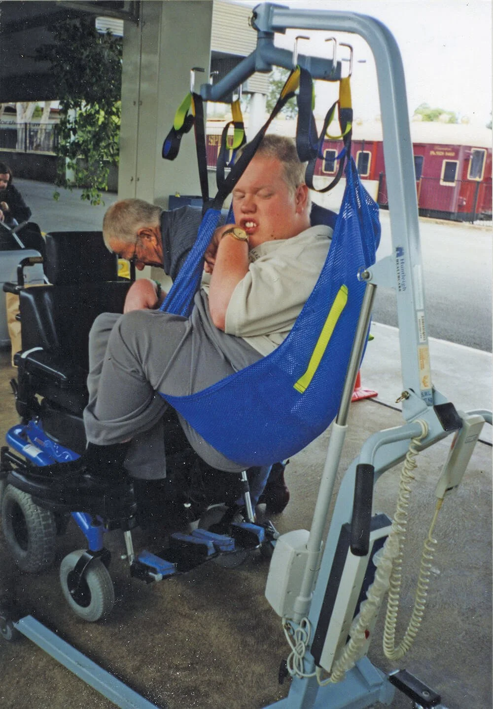 Young man in a wheelchair with a blue safety harness, appearing to be uncomfortable or in pain, next to a hospital bed or medical equipment, with an older woman in the background at a transportation or medical facility.