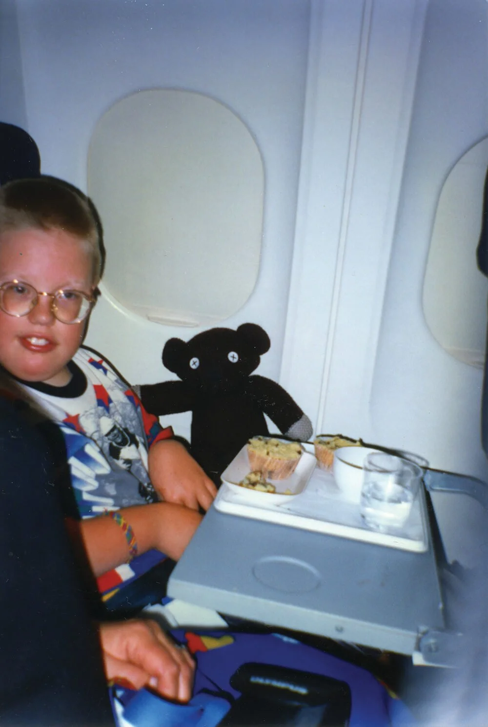 A young boy wearing glasses and a colorful shirt sitting in an airplane seat with a tray table, holding a teddy bear and a meal of cupcakes, cookies, and drinks.