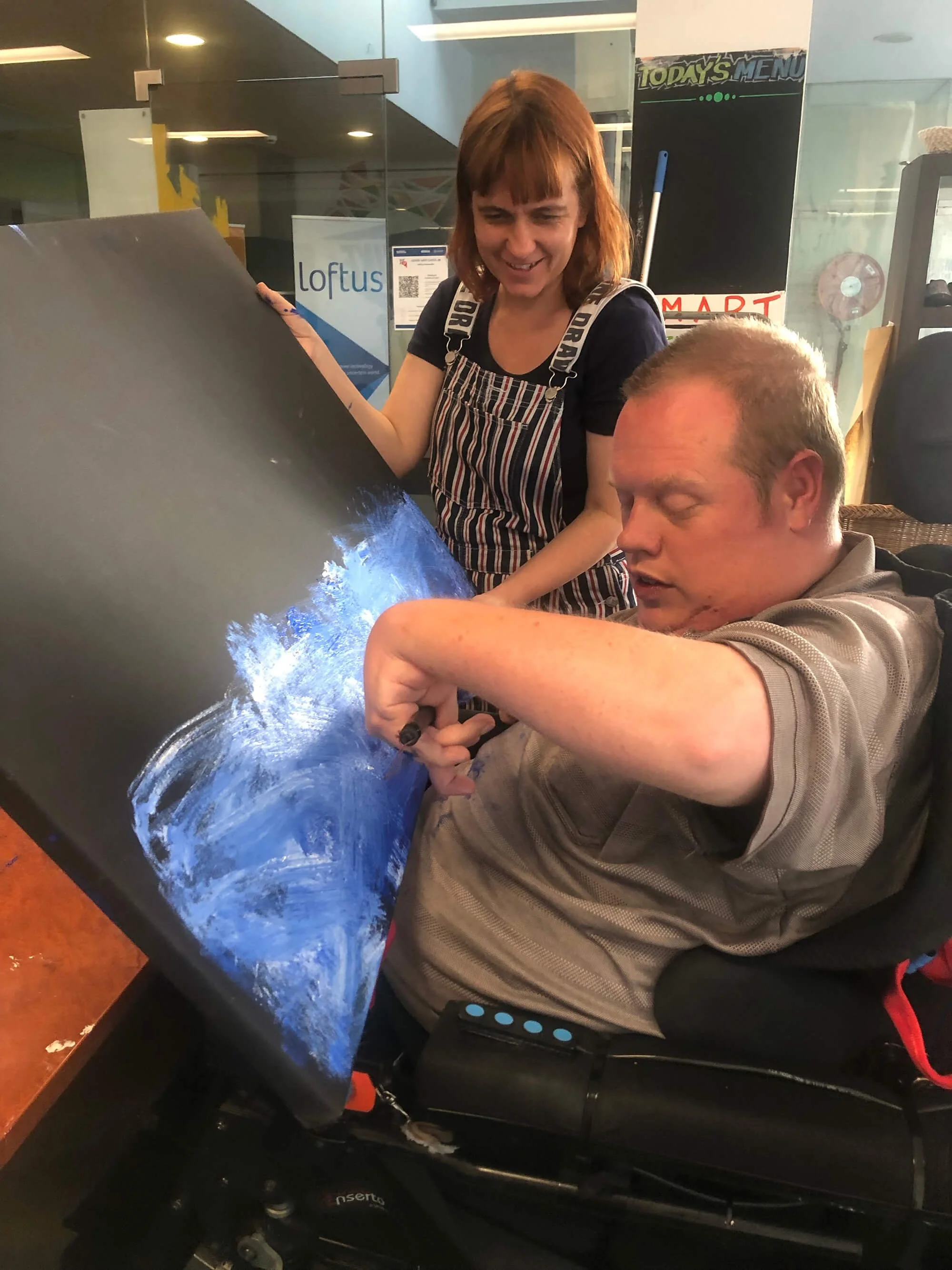 A woman with red hair helping a man in a wheelchair paint a canvas with blue and white colors.