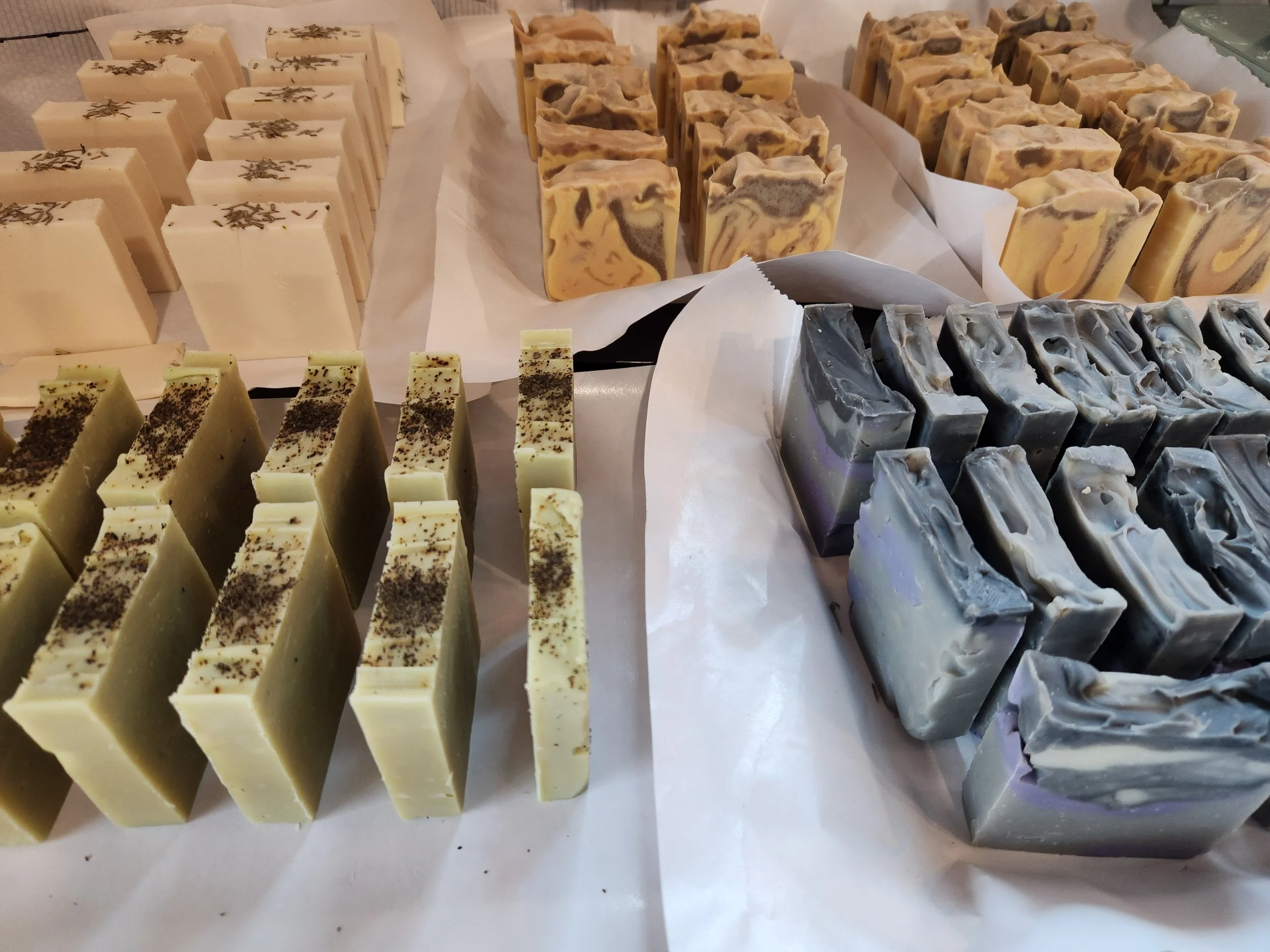 Various handcrafted soap bars arranged on display, including cream-colored with topped with rosemary, marbled beige and brown, tea green topped with green tea leaves, and swirling black and white designs for men's soap.
