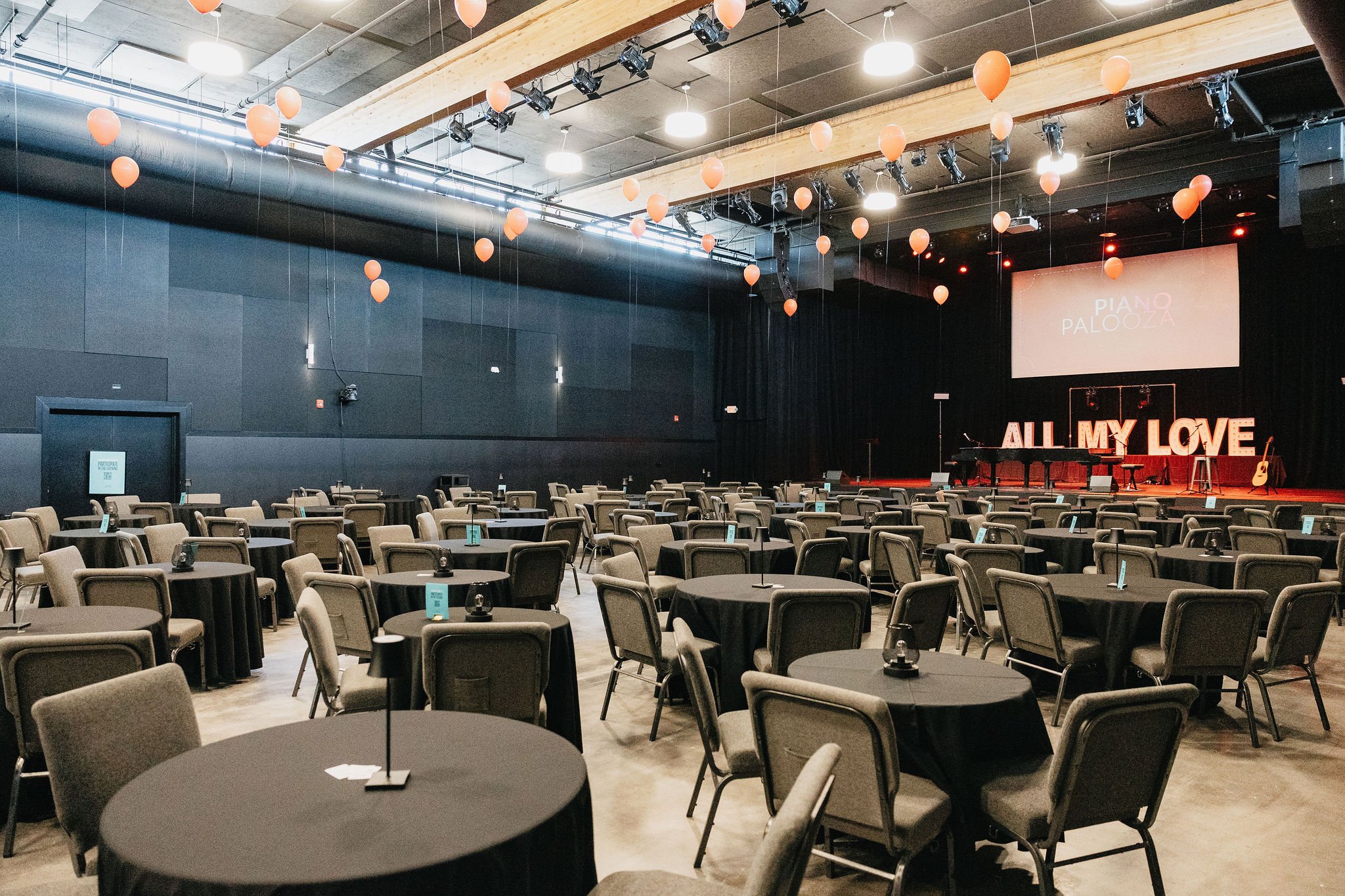 Empty banquet hall or performance space with black round tables, gray chairs, and a stage with large illuminated letters spelling 'All My Love'. Orange balloons are hanging from the ceiling, and there is a screen displaying 'Piano Palooza'.