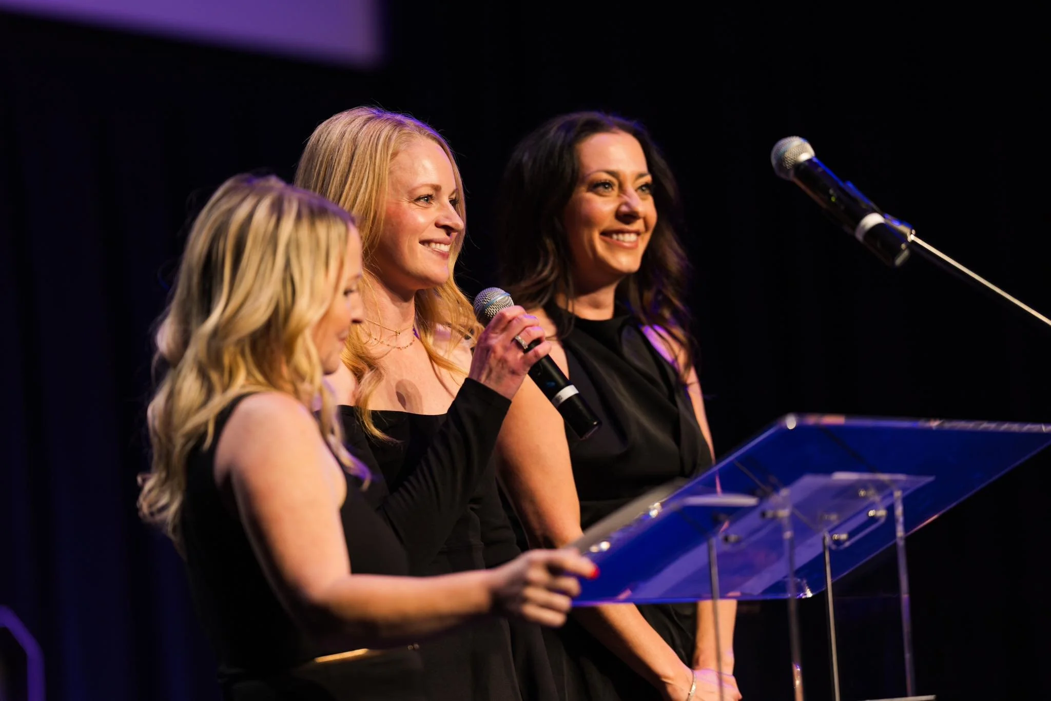 Three women standing at a podium with microphones, smiling, during an event or presentation, dressed in black at Zootown Event Center, raising money for VEO Guardians