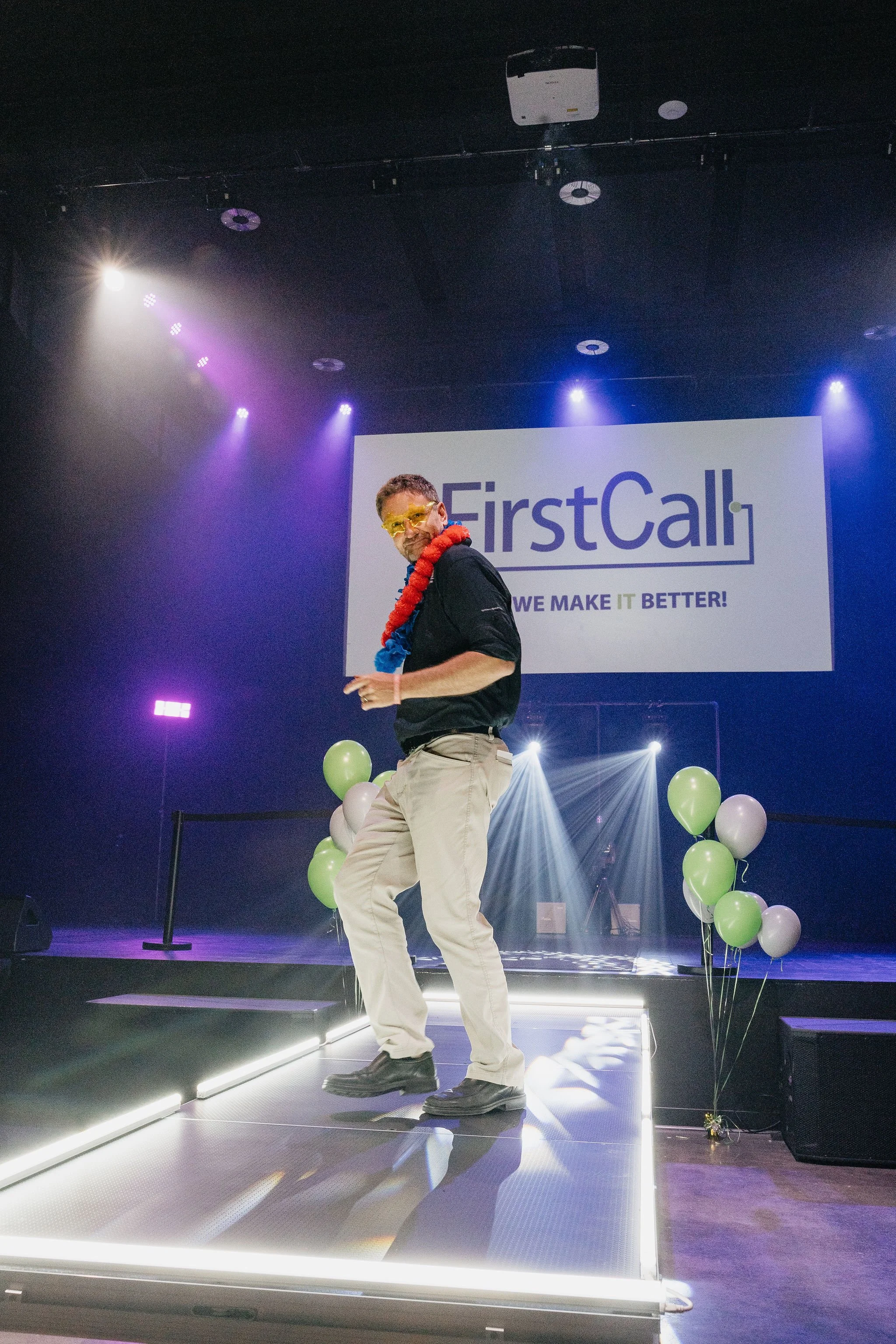 A man on a decorated stage with balloons, wearing sunglasses and a colorful necklace, at a FirstCall corporate event in Missoula at Zootown Event Center.