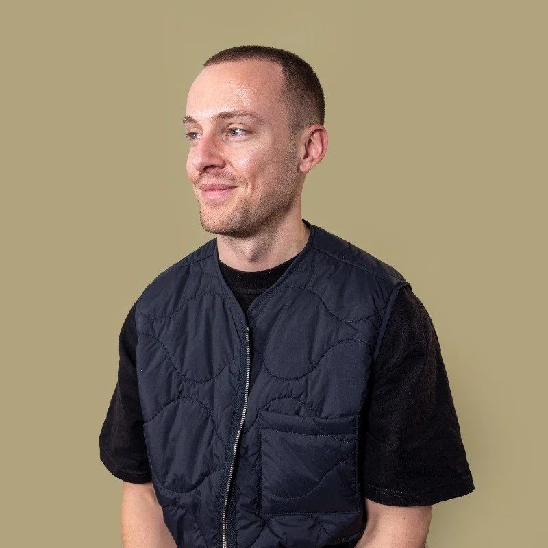 A man with short hair and a slight smile, wearing a black shirt with a sleeveless dark blue quilted vest, standing against a plain beige background.