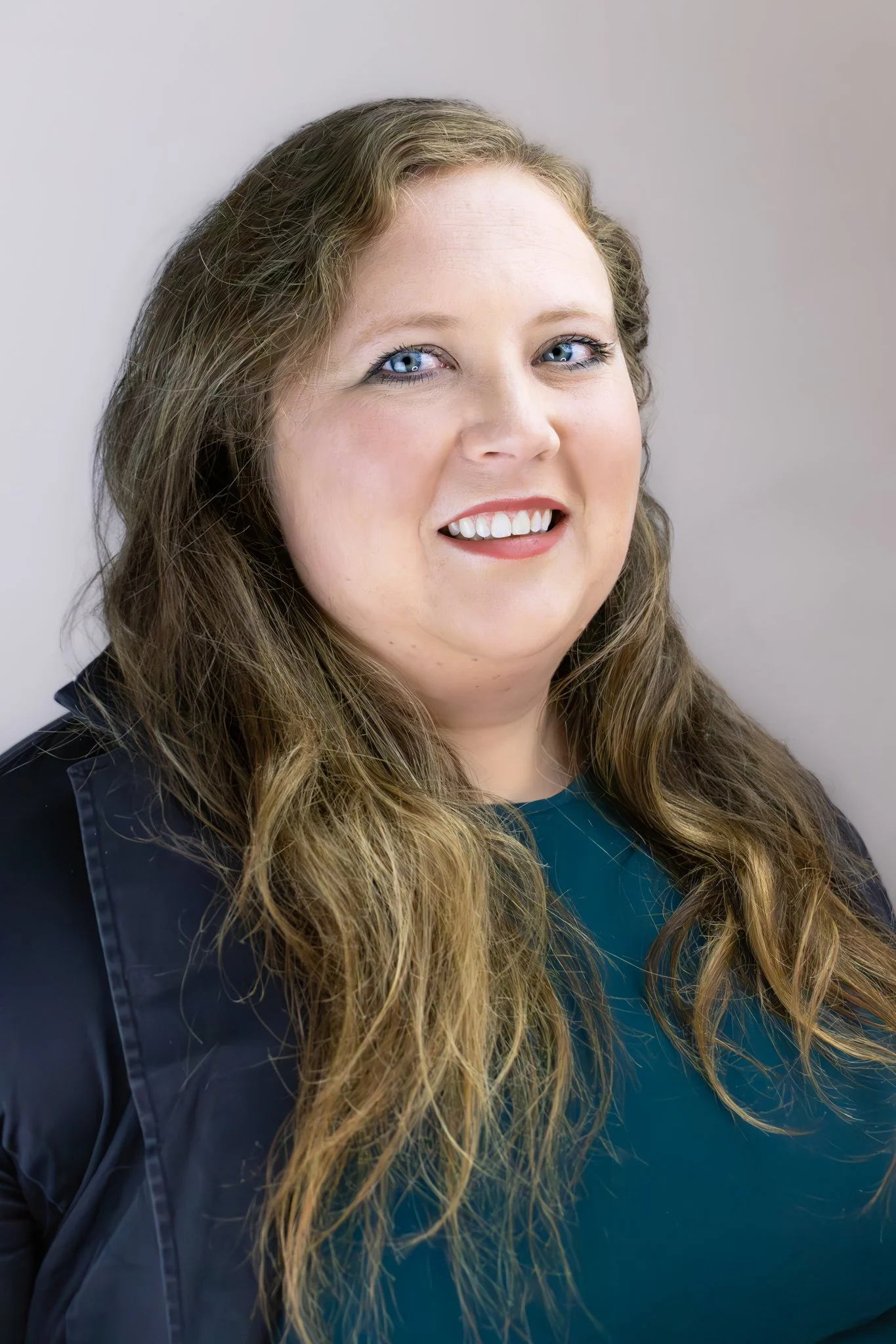 Close-up portrait of a woman with long wavy hair, blue eyes, wearing a teal top and black jacket, smiling.