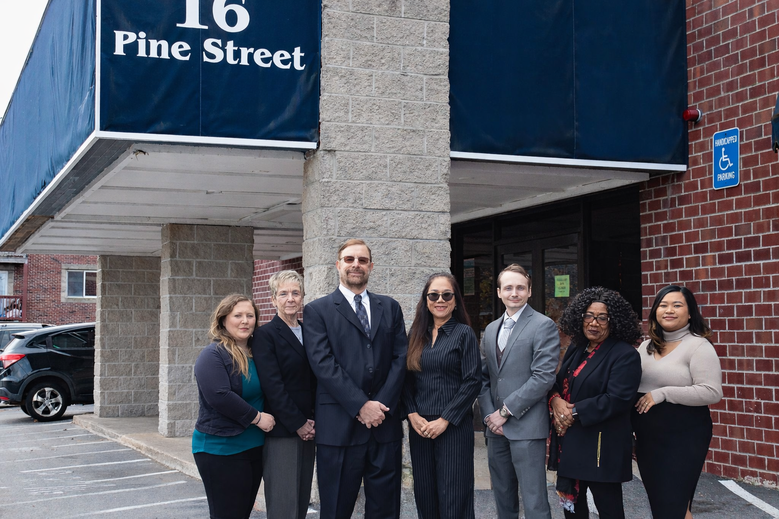 Group of seven diverse professionals standing in front of a building with a sign that reads '16 Pine Street'. The group includes men and women dressed in business attire, smiling and posing for the photo. There are parked cars and a handicap parking sign visible.