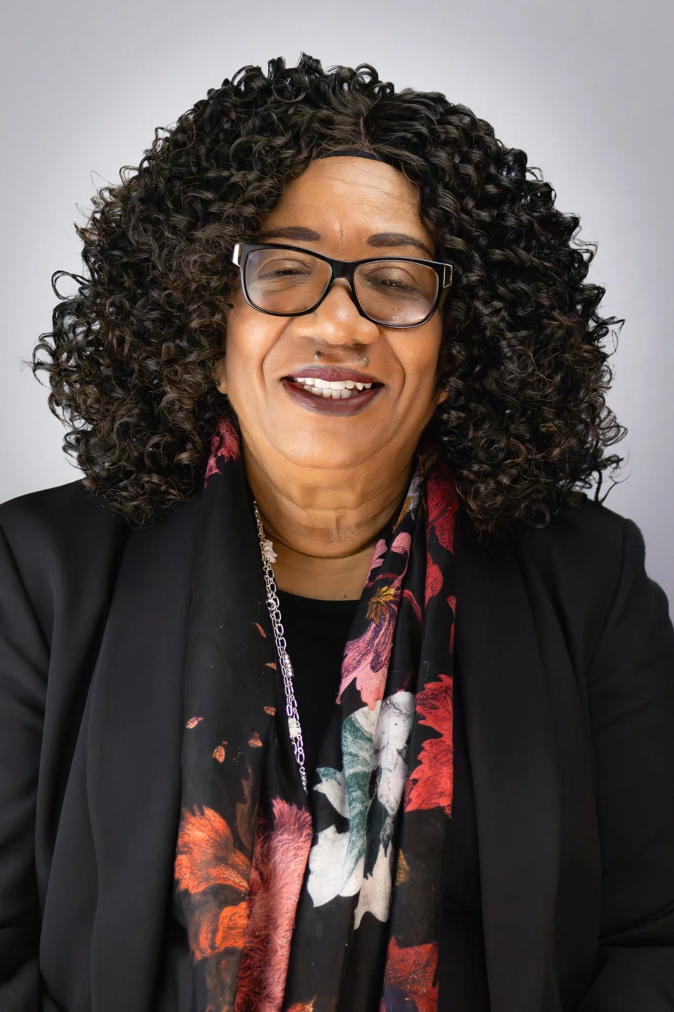 A woman with curly black hair, glasses, and dark lipstick smiling, wearing a black blazer and a floral scarf, against a plain light background.
