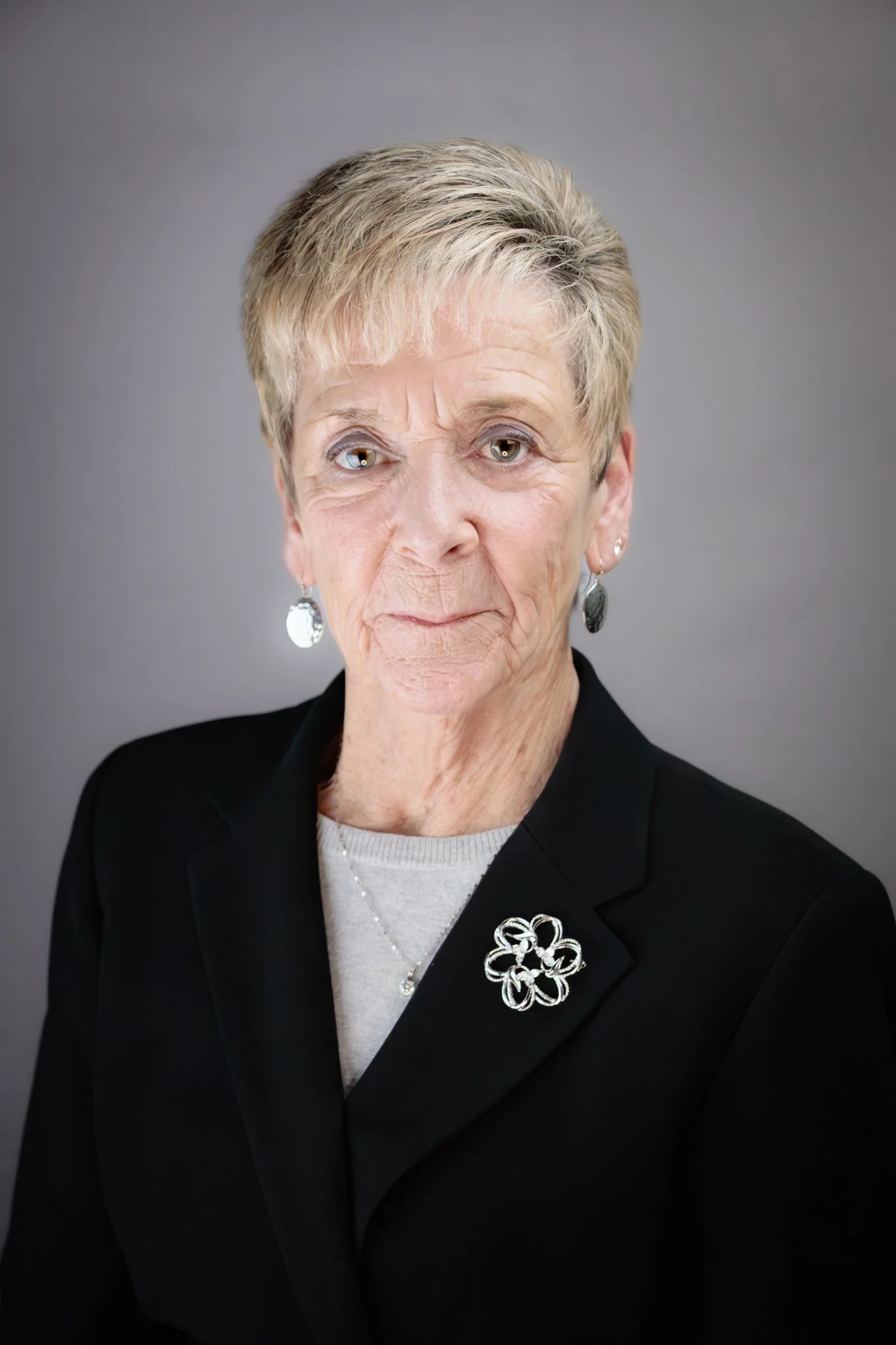 An elderly woman with short, light-colored hair, wearing a black blazer with a floral brooch and earrings, against a gray background.