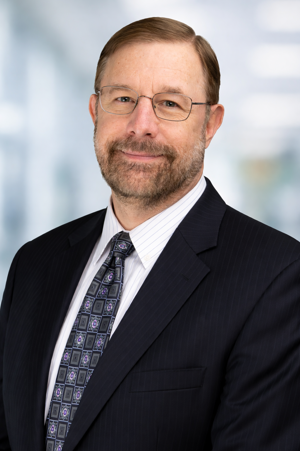 Portrait of a middle-aged man with glasses and a beard, wearing a dark business suit, white shirt, and patterned tie, smiling in front of a blurred office background.