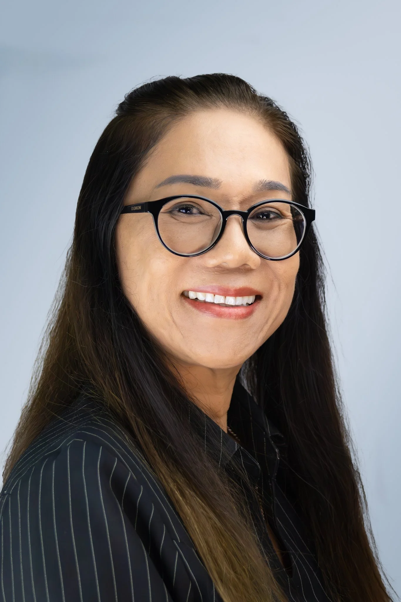 A smiling woman with long dark hair, wearing glasses and a dark pinstripe jacket, against a light blue background.