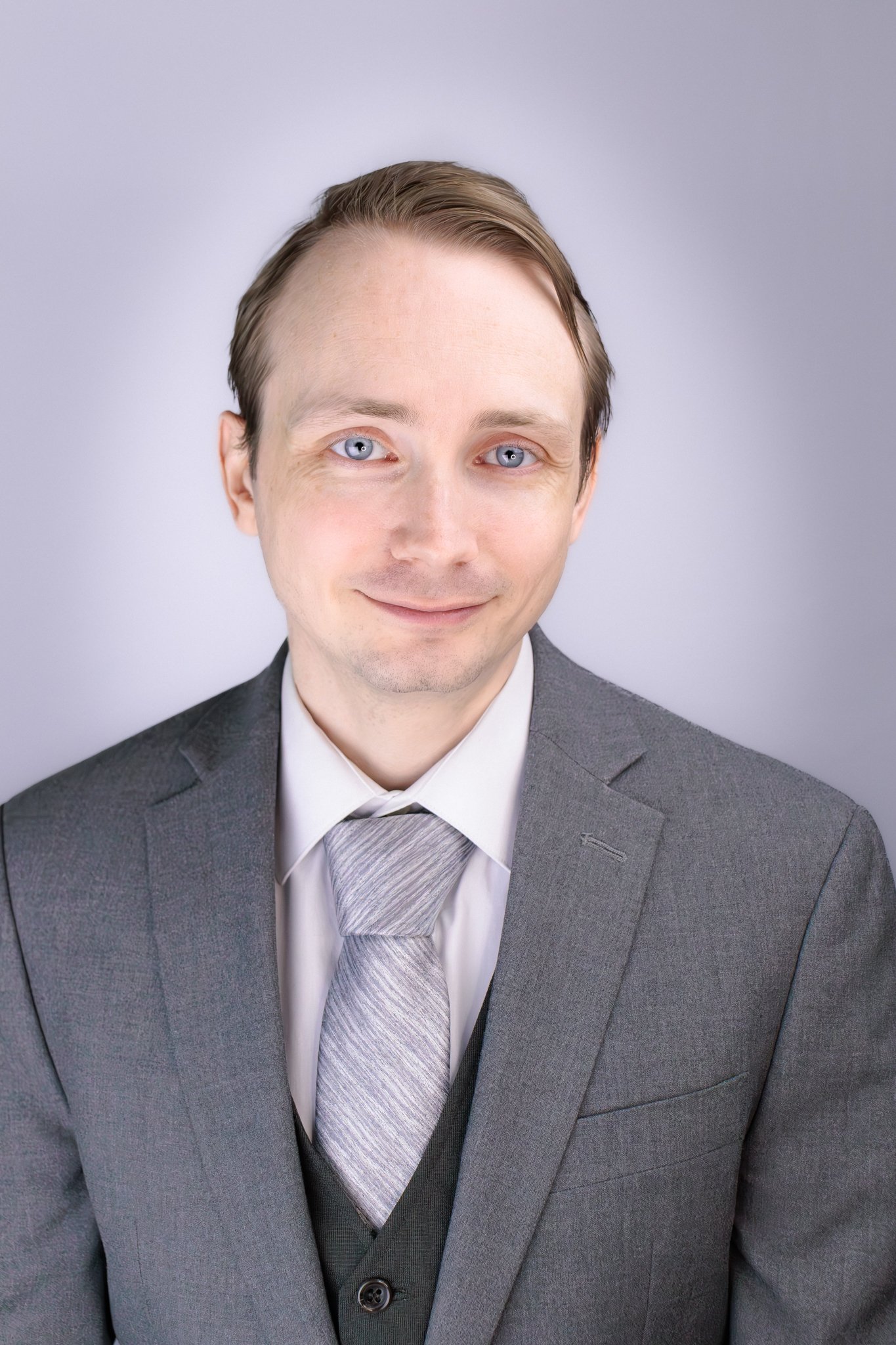 Portrait of a young man in a grey suit, white shirt, and patterned tie, smiling with a plain background.