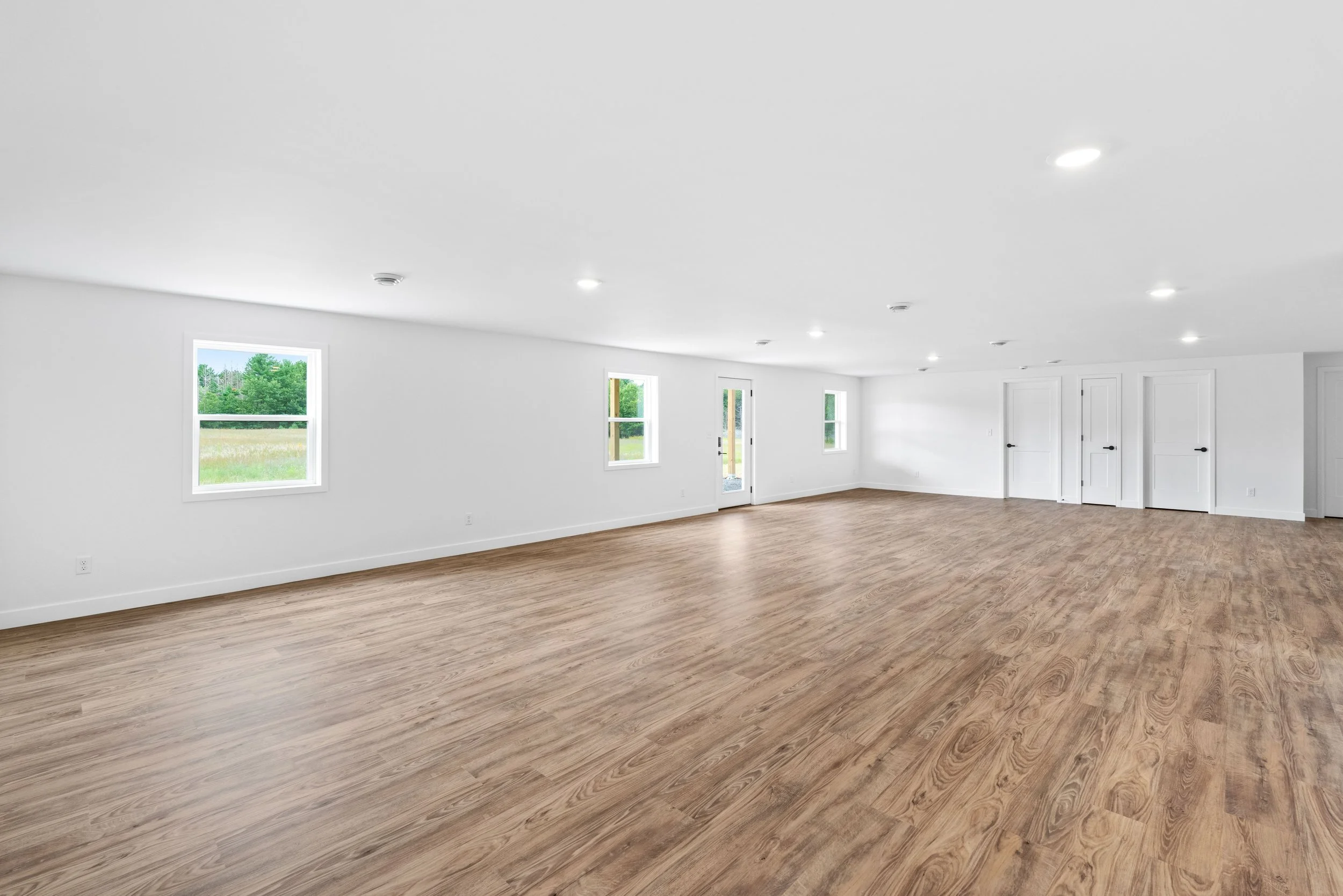 Empty spacious room with white walls, multiple windows, wood flooring, and ceiling lights.