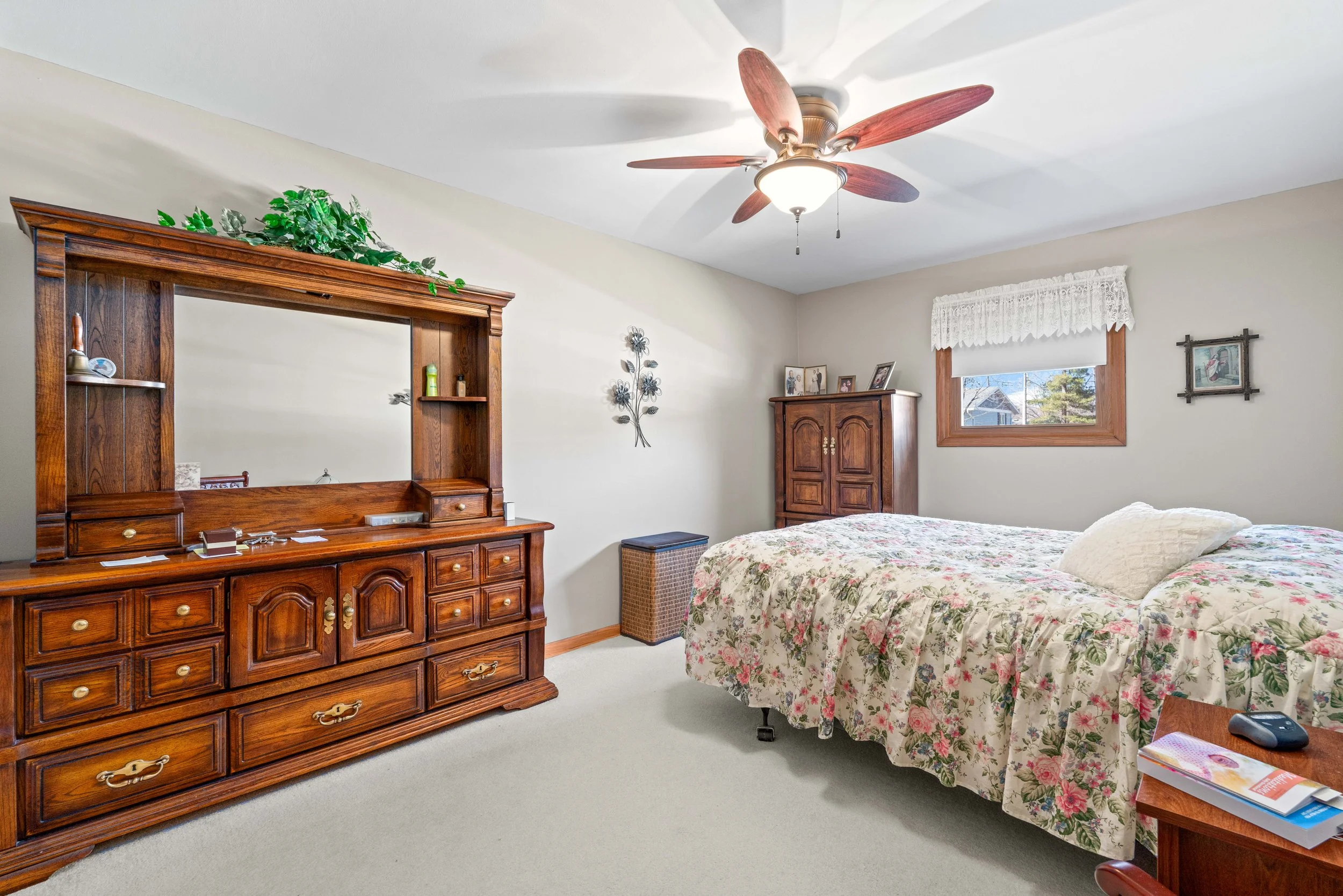 A bedroom with a wooden dresser, a wall-mounted mirror, a bed with a floral quilt, a wooden wardrobe, a small window with curtains, and a ceiling fan.