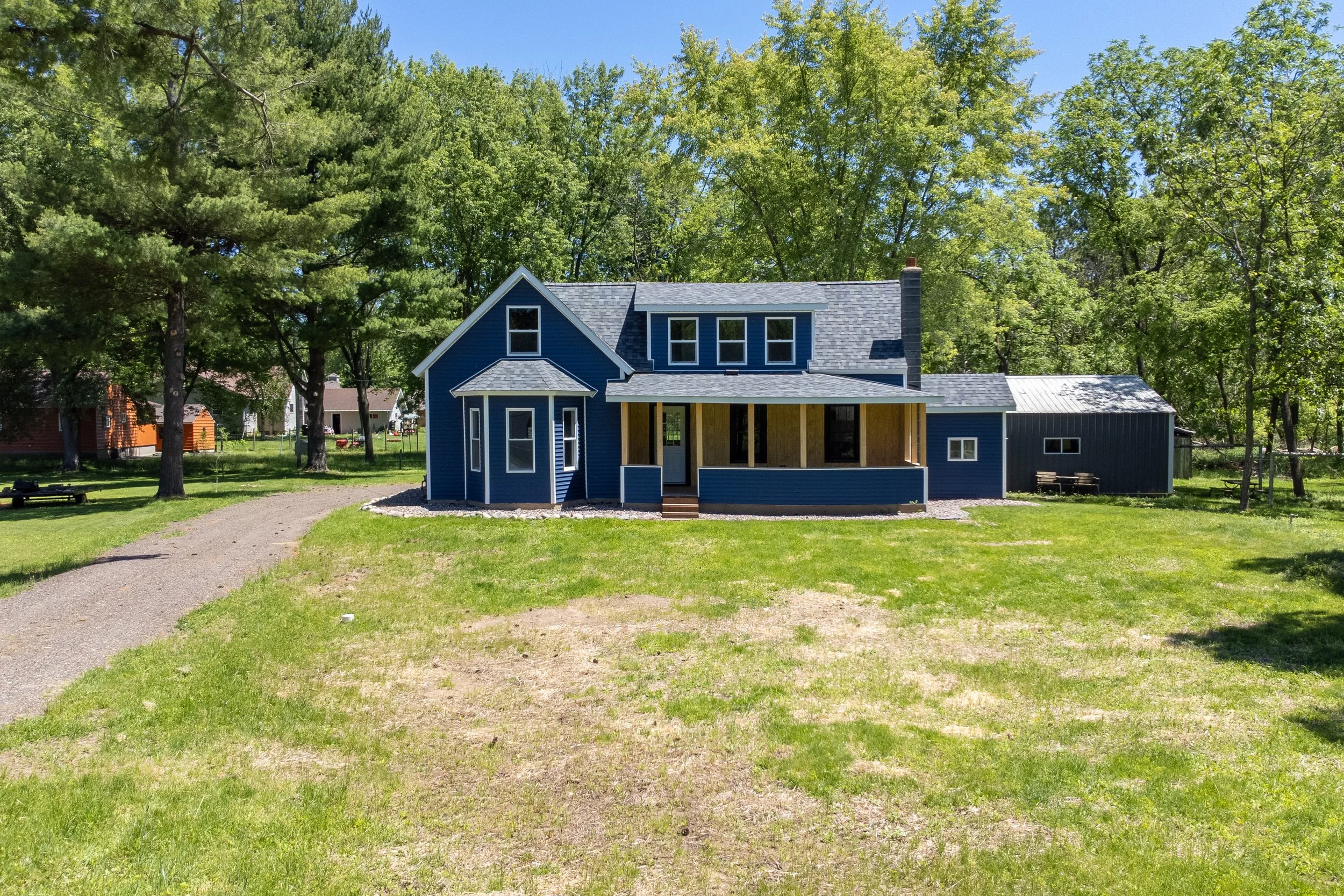 A blue house under construction in a grassy yard with tall trees in the background, sunny sky, and a gravel pathway leading to the house.