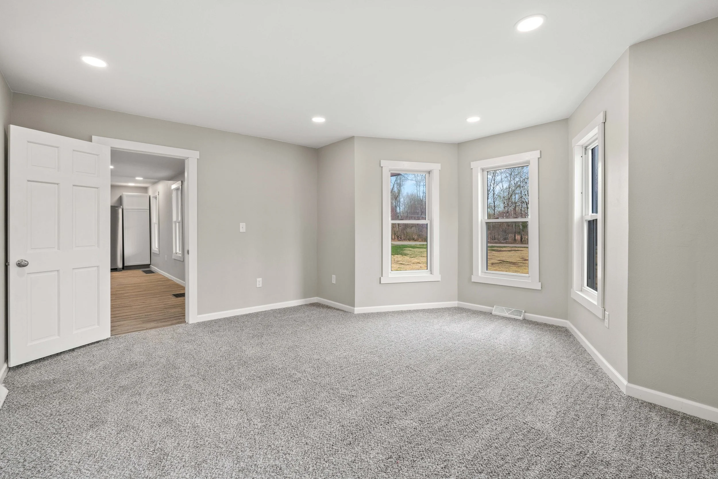 Empty living room with light gray walls, three large windows, gray carpet, and a doorway leading to a kitchen area with wood flooring and stainless steel refrigerator.