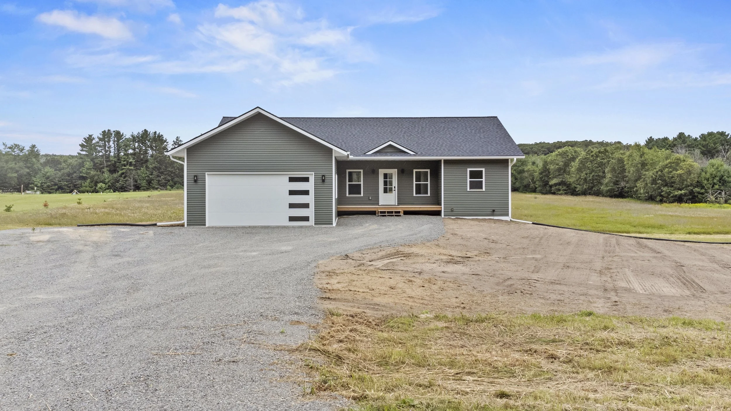 Newly constructed gray house with a white garage door, front porch, and surrounded by open land with a gravel driveway and some grass.