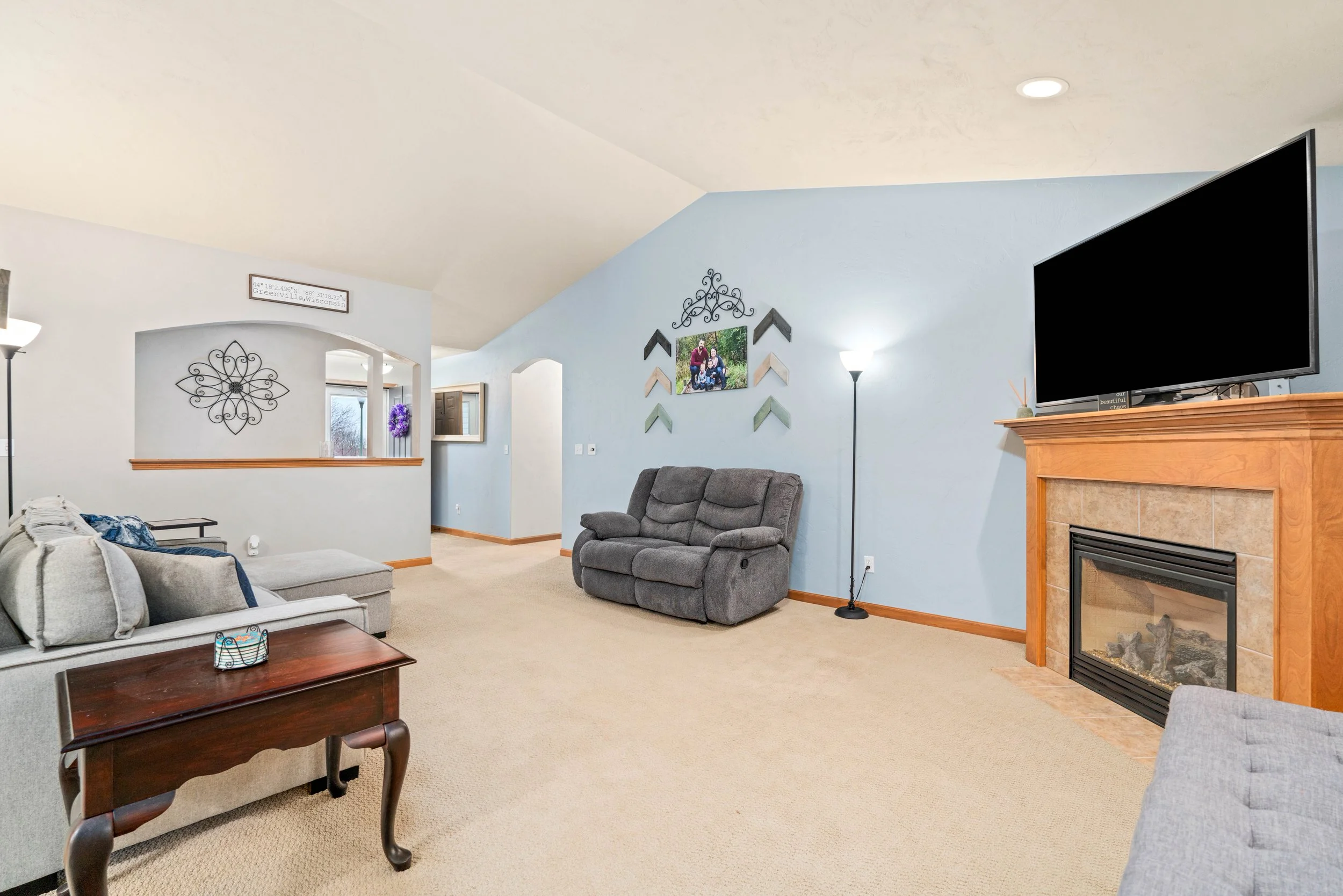 Living room with beige carpet, a gray recliner sofa, a beige sectional couch, a wooden coffee table, a gray armchair, a coastal-themed wall decor, a fireplace with a black TV on top, and a floor lamp, with a decorative sign and a picture frame on the