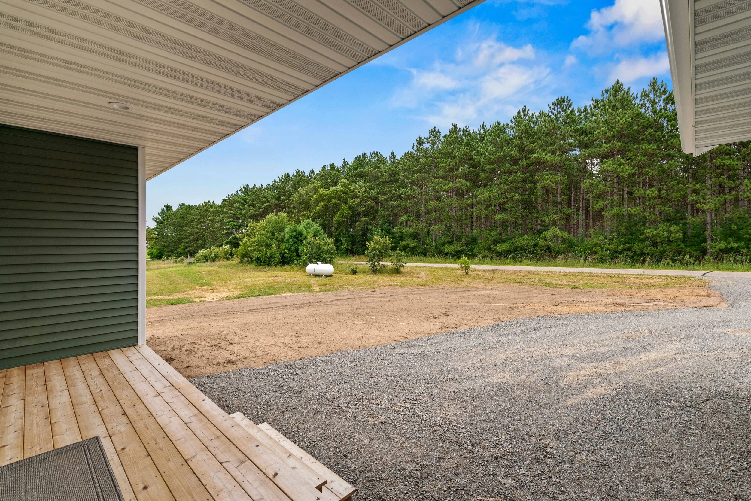 View from a house porch showing a gravel driveway, grassy yard, and a forested area with tall trees under a partly cloudy sky.