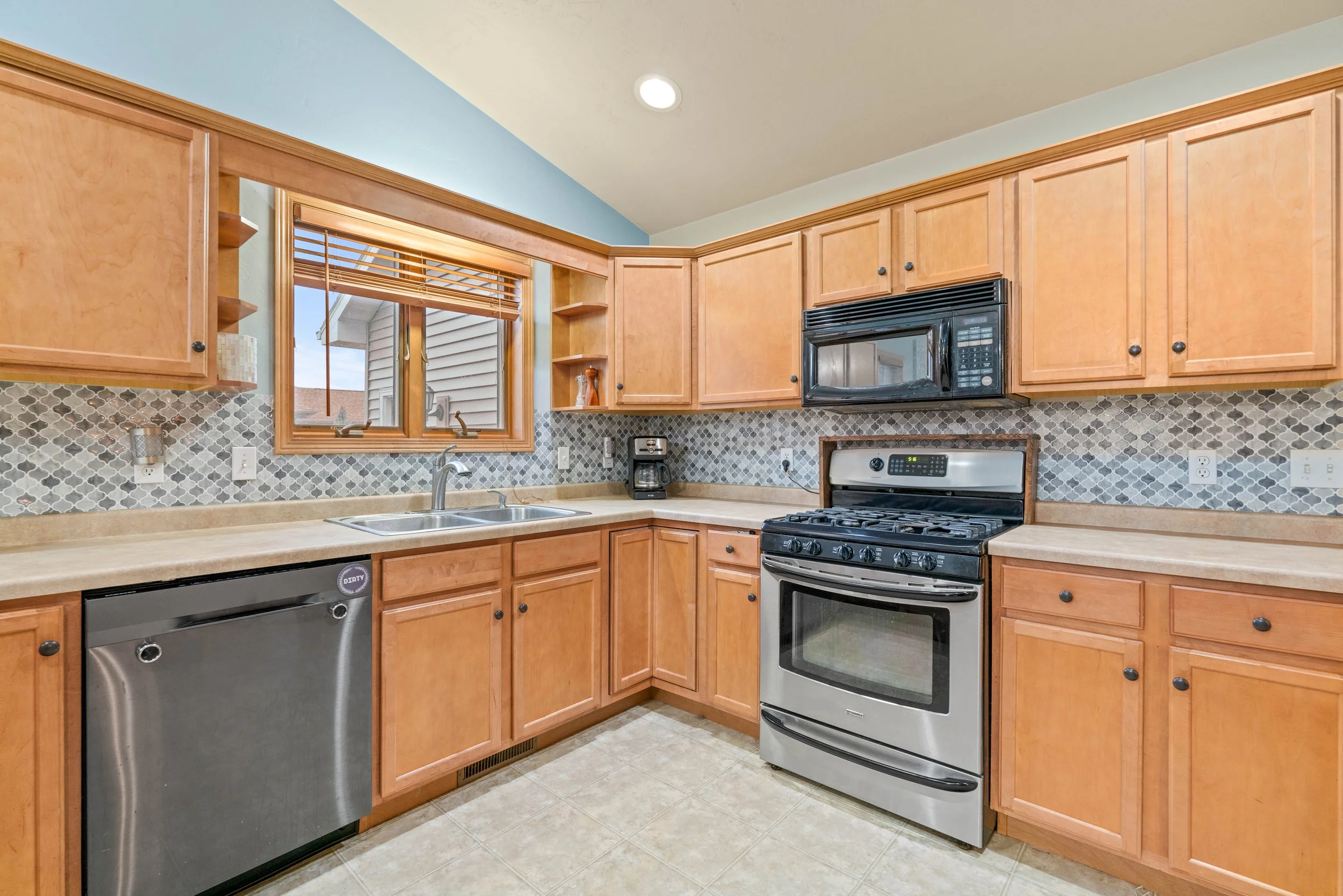 Kitchen with wooden cabinets, a window over the sink, black microwave, stainless steel stove and dishwasher, beige countertops, gray patterned backsplash, and tiled floor.