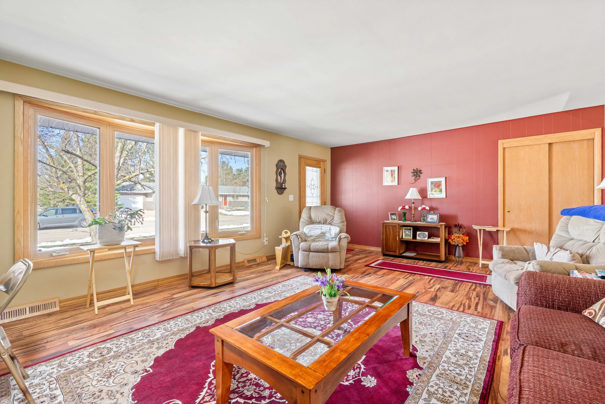 Living room with large windows, beige and red walls, hardwood floors, and various furniture including sofas, armchairs, a coffee table with a flower pot, and a wooden cabinet. Decor includes framed pictures, lamps, and decorative items.