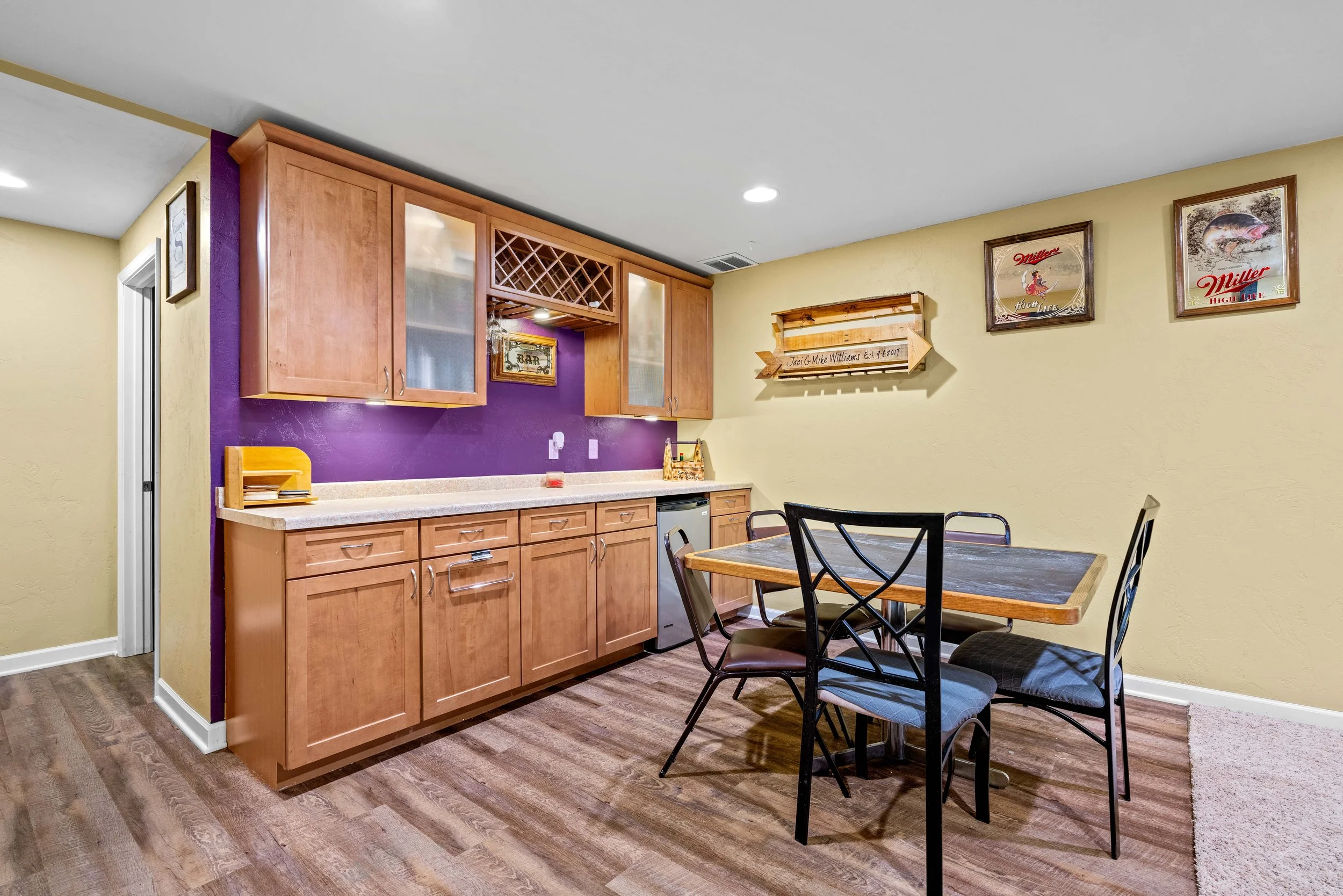 Kitchen area with wooden cabinets, a small refrigerator, a colorful wall with memorial and decor items, and a dining table with four black chairs.