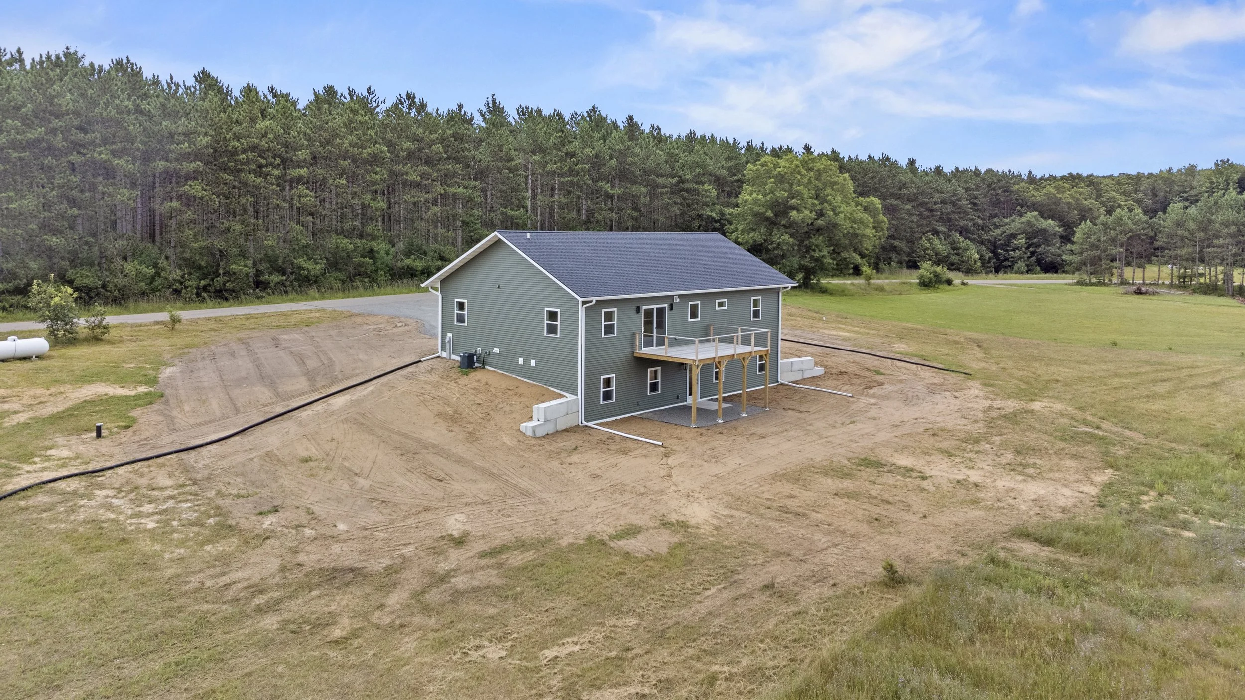 A newly built house with green siding and a dark gray roof on a large, open lot with a backyard deck overlooking a grassy field and woods in the background.