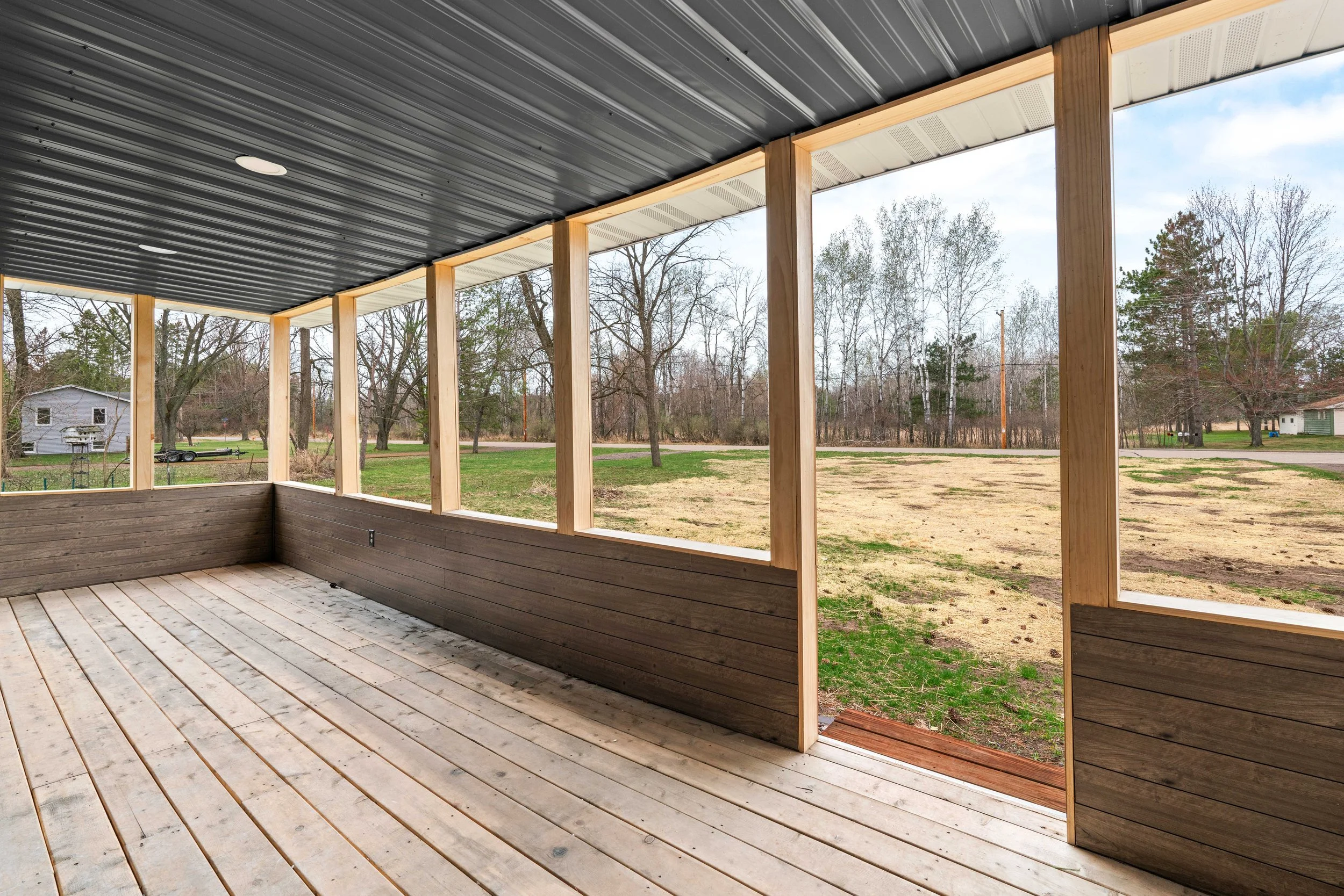 A partially enclosed wooden porch with a metal roof, overlooking a grassy yard with trees and a distant house.