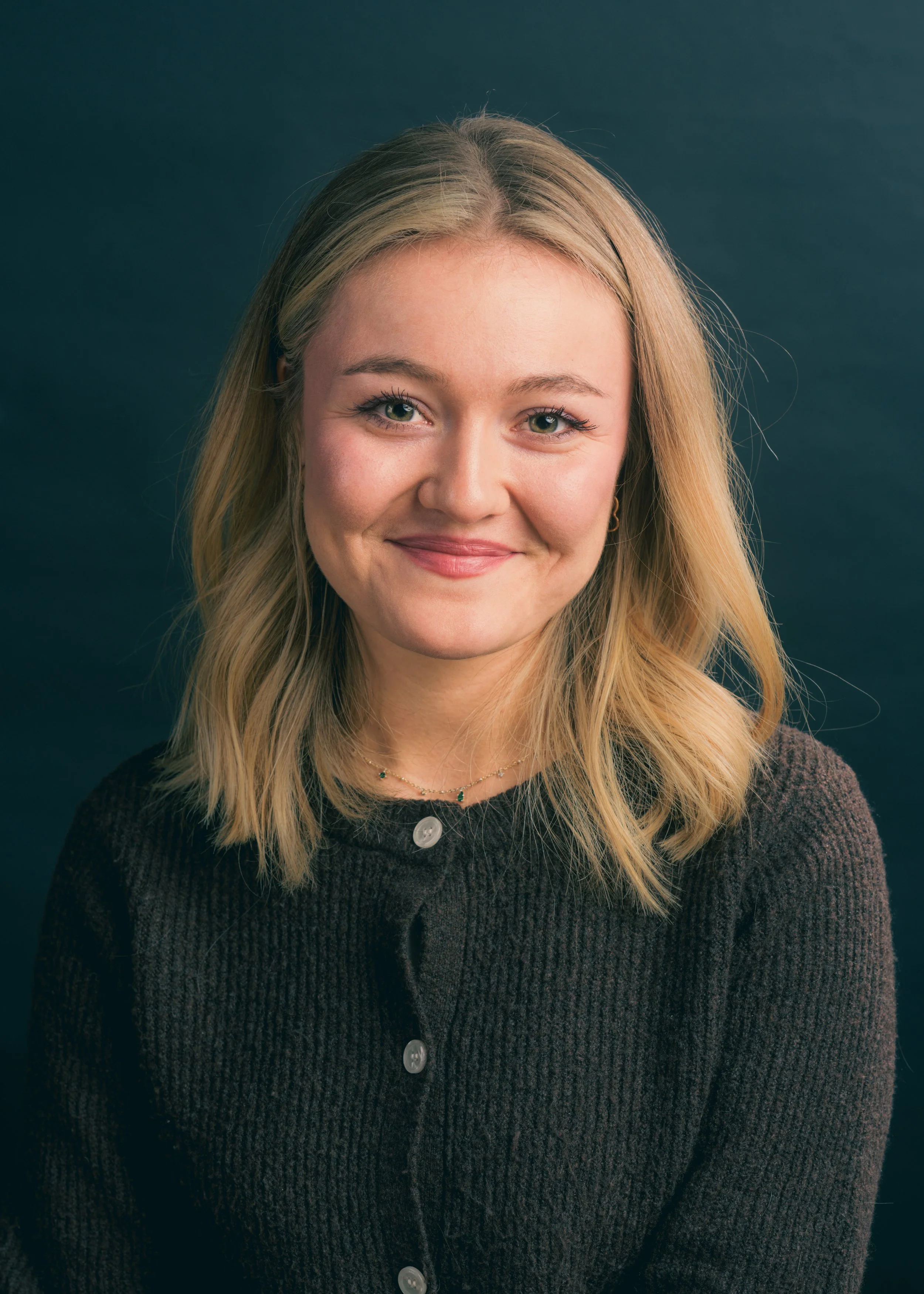 A woman with blonde hair, green eyes, and a light complexion, smiling in front of a dark background, wearing a dark knit sweater and a delicate necklace.