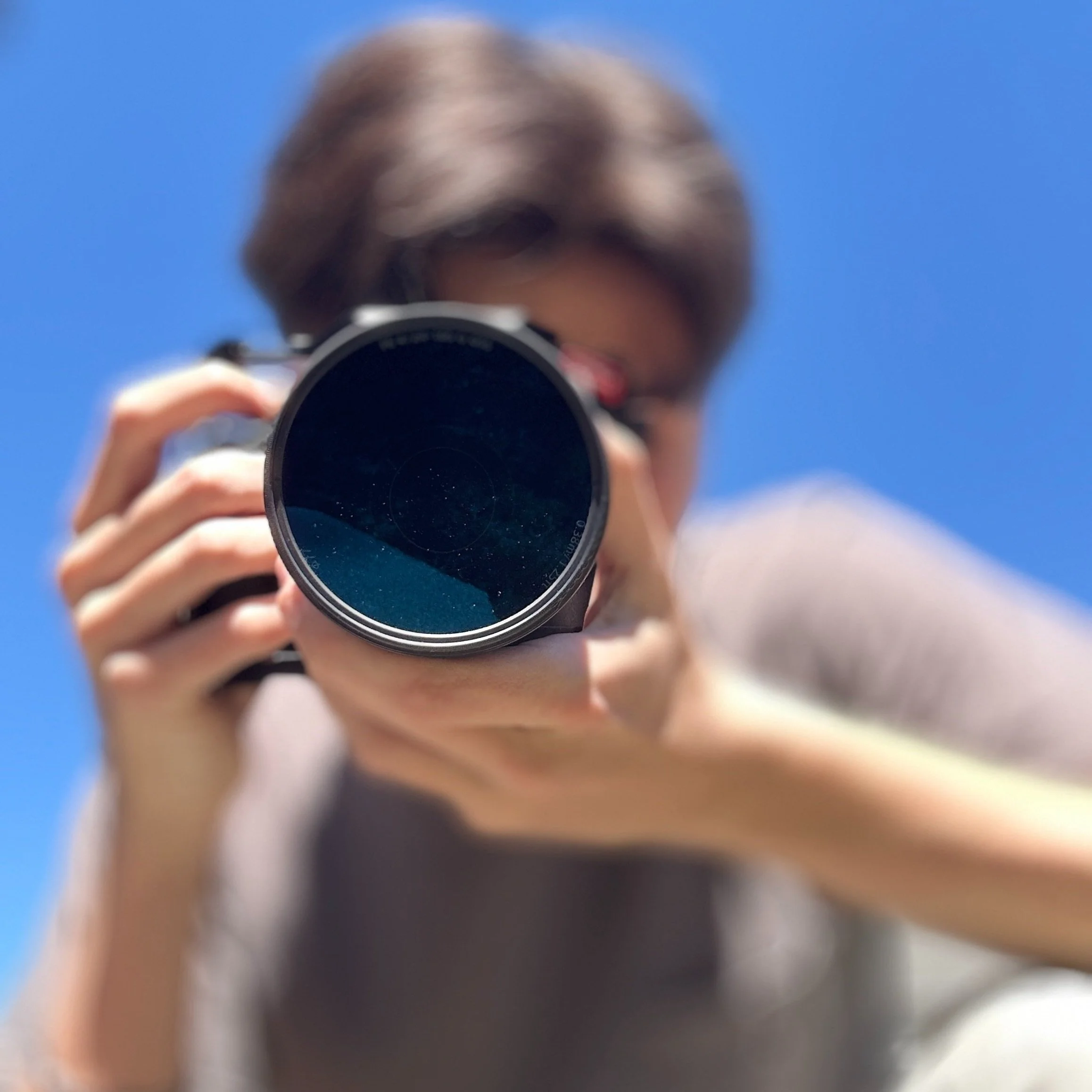 Person holding a camera with a large lens, pointing the lens towards the viewer, blue sky in the background.