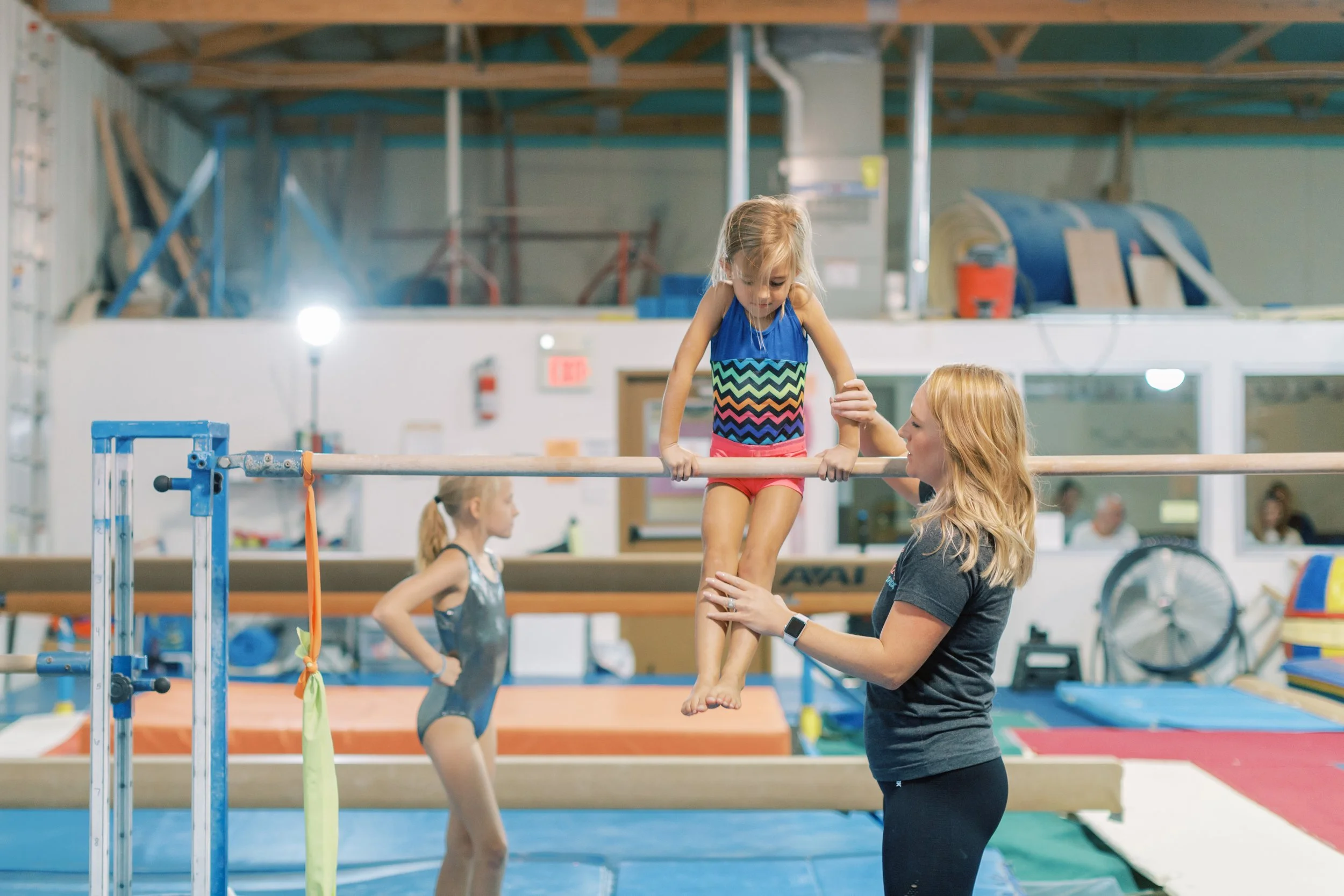 A young girl practicing gymnastics on parallel bars with the assistance of a coach in a gymnasium environment. Another child waits in the background.