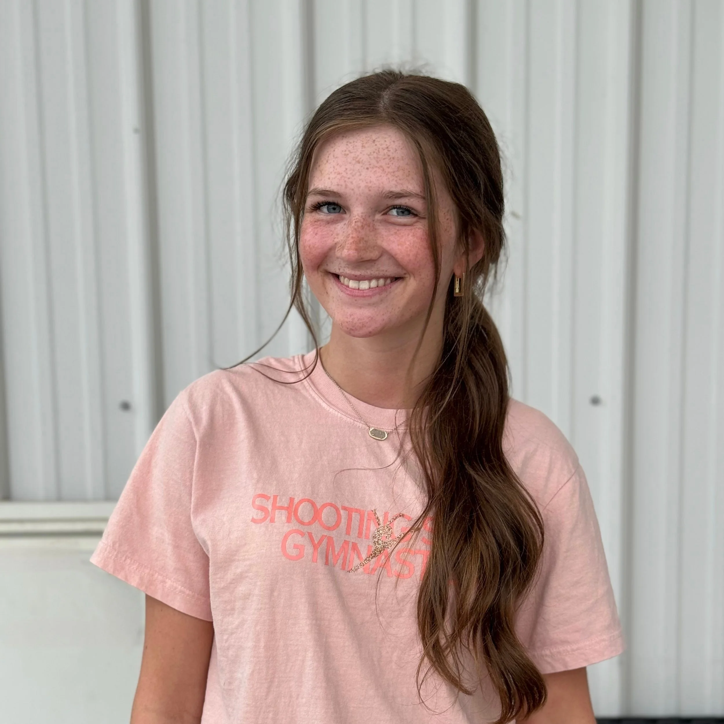 Young woman smiling in a pink t-shirt with text "Shooting Star Gymnast" standing in front of a white metal wall.