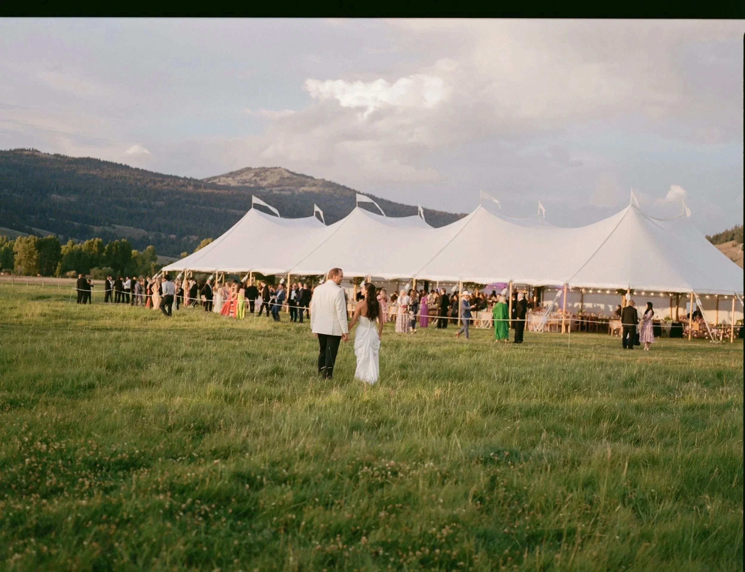 A wedding celebration taking place outdoors on a grassy field with a large white tent in the background, surrounded by mountains and a partly cloudy sky.