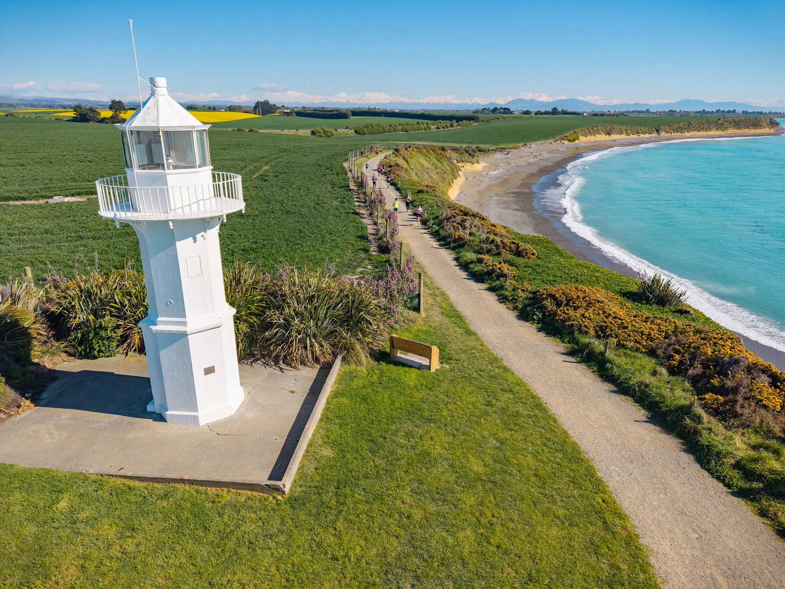 A white lighthouse on a grassy area by the coast with a walking trail, overlooking the ocean and a sandy beach, with green fields and mountains in the background.