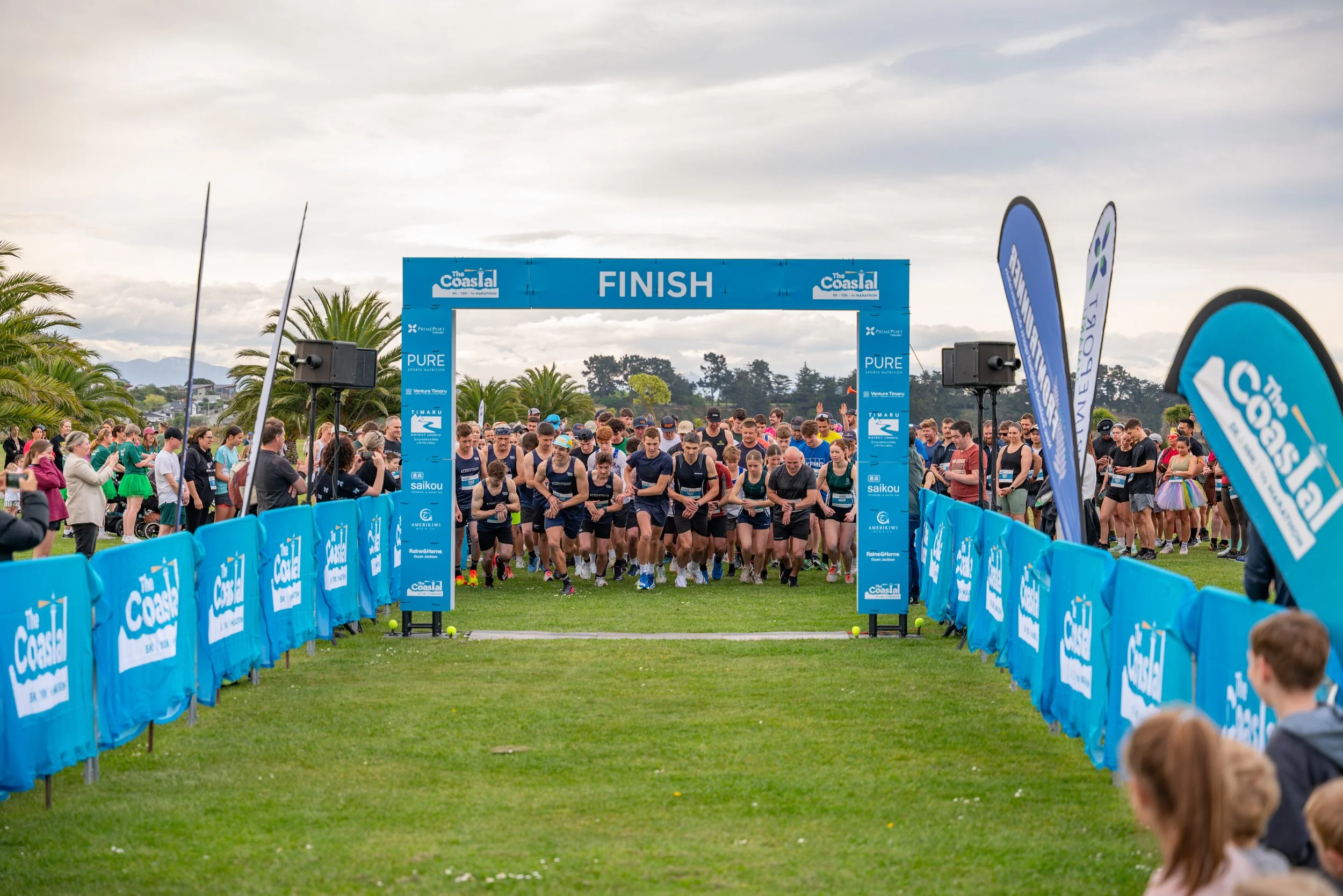 Group of runners at the starting line of a race, with a blue finish line banner overhead. Spectators and event staff are on the sides, and flags are visible. The scene is outdoors with palm trees and cloudy sky in the background.