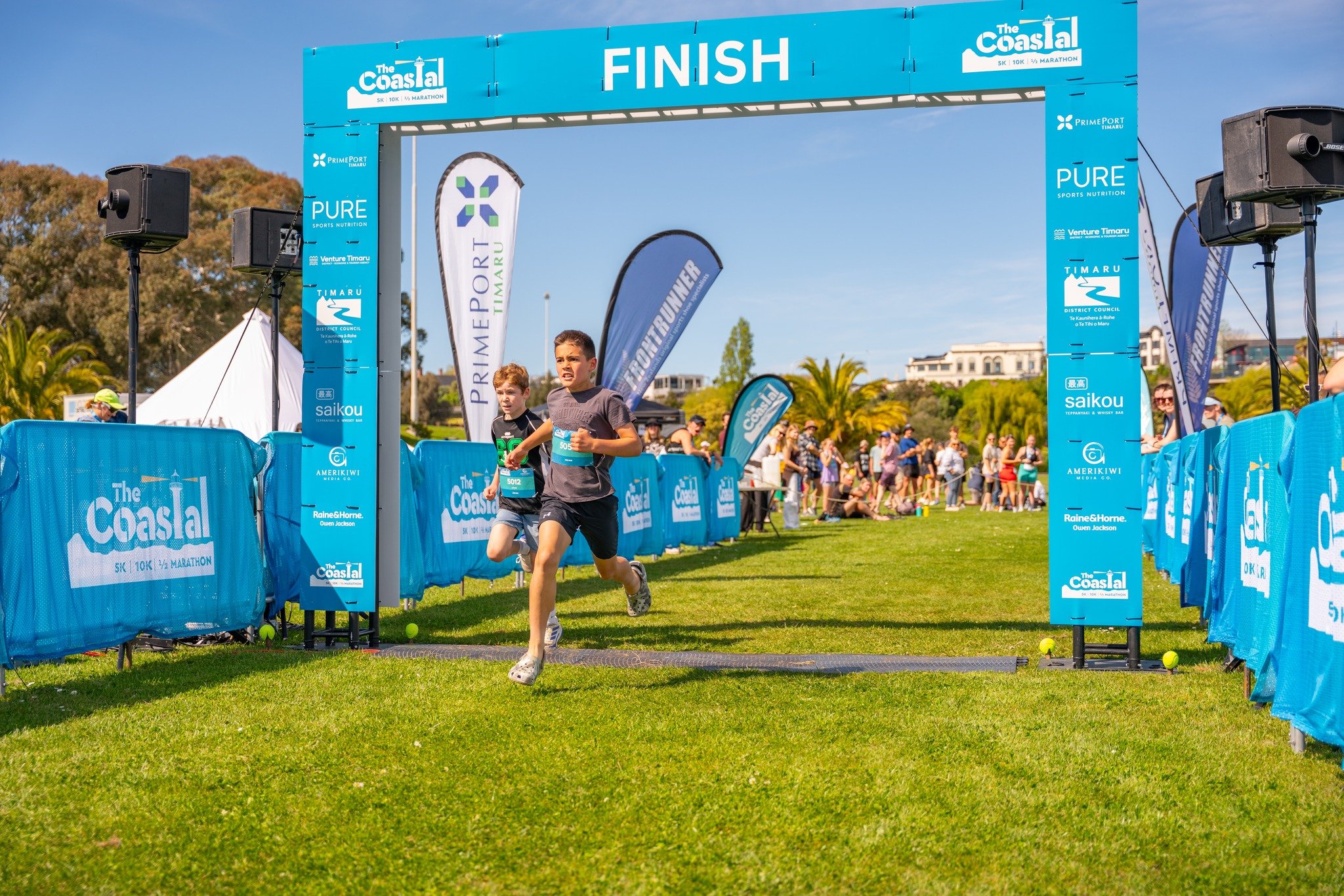 Children finishing a race under a blue finish line banner with the words "The Coastal 5K 10K Half Marathon" on it, with spectators and event flags in the background on a sunny day.