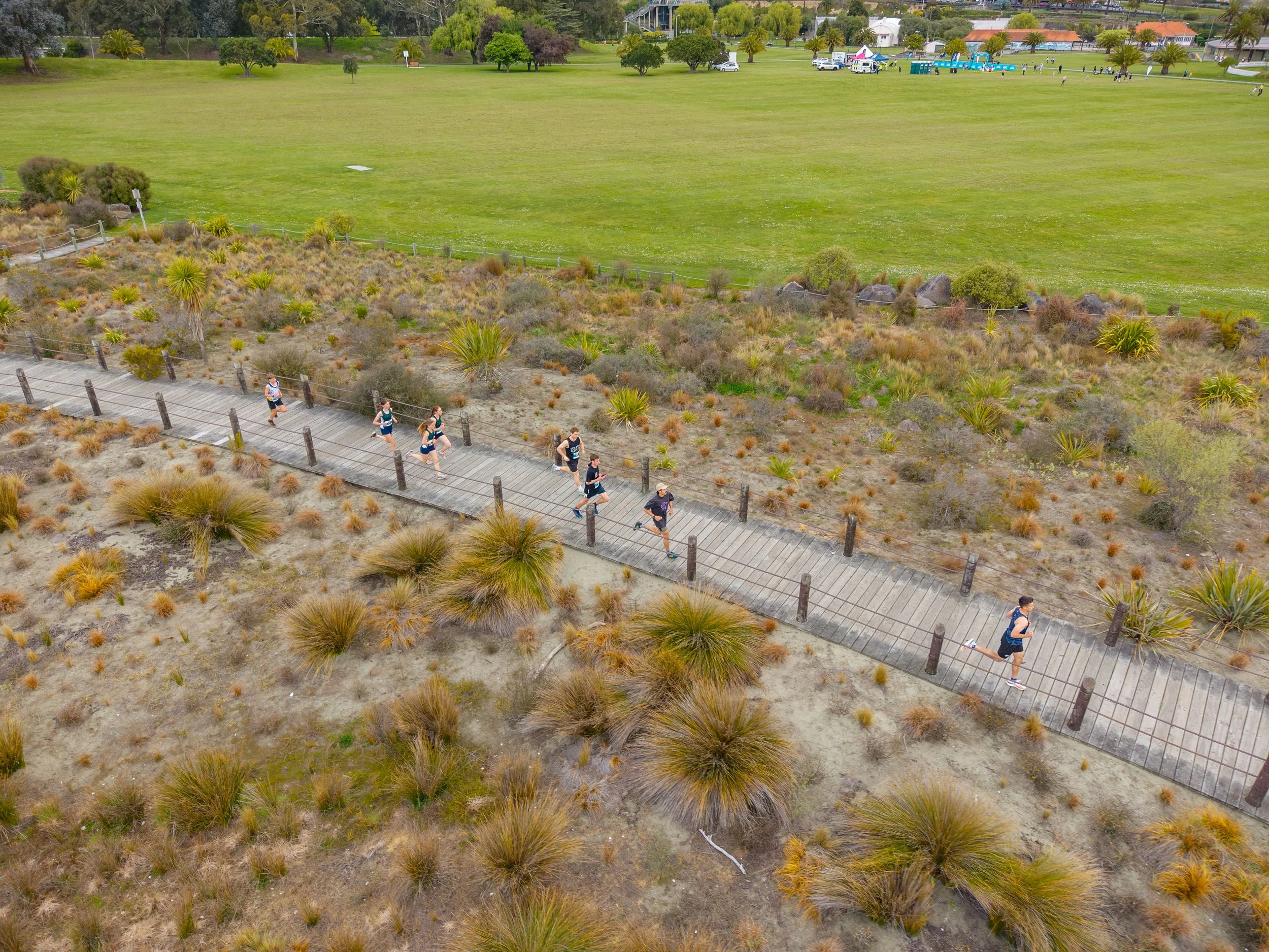 Aerial view of a group of runners jogging on a wooden pathway through a desert landscape with sparse vegetation, bushes, and grasses. There is a large grassy area and some buildings in the background.